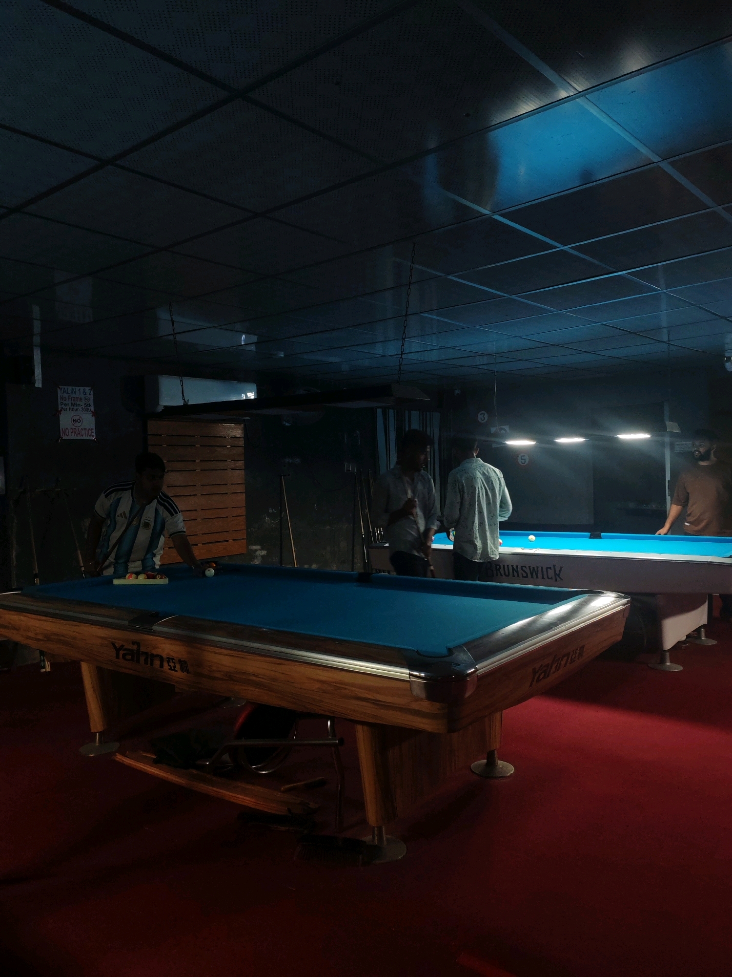 A dimly lit indoor pool hall in Cumilla, Bangladesh features multiple billiards tables, three men, and a sign. In the foreground, a pool table with a wooden frame and blue felt is visible. To the left, a man wearing a blue and white striped soccer jersey is standing over the table, holding a pool cue and looking down at the balls. A rack of colored pool balls sits on the table. In the background, two other men are standing near a second pool table. One man, wearing a light-colored shirt, is holding a cue, and the other man, wearing a patterned shirt, is facing away from the camera. A third man in a brown shirt is standing to the right, looking towards the pool tables. A sign with white text on a black background is mounted on the wall behind the first man, reading "YALIN 1 & 2 NO FRAME Per Hour- 5tk Per Hour- 90tk NO PRACTICE". The ceiling is a dark, tiled surface with visible light fixtures and ductwork. The floor is covered in red carpeting.