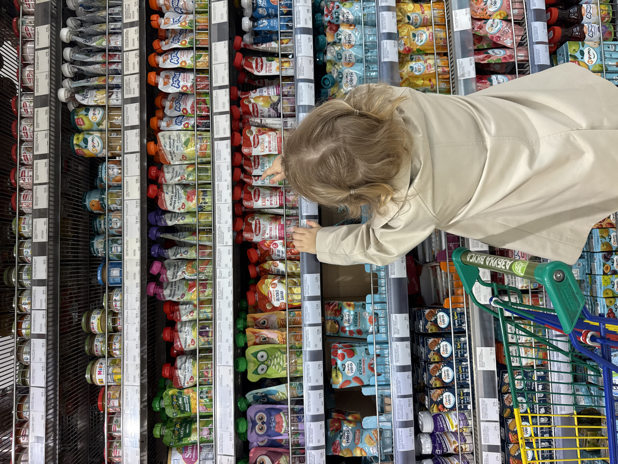 The image, taken from an overhead perspective and rotated 90 degrees clockwise, shows a young child from behind, reaching for products on a supermarket shelf. The child has blonde hair tied in two small pigtails and is wearing a light beige jacket. Their left hand is extended towards a row of baby food pouches.
The scene is an aisle in a grocery store, specifically dedicated to baby and children's food. The shelves are packed with a variety of products including squeezable fruit and vegetable puree pouches, glass jars of baby food, and baby snacks like biscuits and fruit pieces. Prominent brands visible are ФрутоНяня (FrutoNyanya), Агуша (Agusha), Semper, Hipp, pymo, and Gipopo. Price tags with Russian Ruble denominations (e.g., "229.-", "75.90") are visible below many items.
A green-handled shopping cart, with blue and yellow sections, is positioned to the right of the child. The cart's handle clearly displays "АЗБУКА ВКУСА" (Azbuka Vkusa), identifying the supermarket chain, which, along with the Russian text on product labels such as "КАША" (porridge), "ФРУКТОВОЕ ПЮРЕ" (fruit puree), "ЯБЛОКО" (apple), and "ПЕЧЕНЬЕ" (biscuits), confirms the location as Moscow, Russia. The activity depicted is a child selecting items during a grocery shopping trip. The lighting is artificial, consistent with an indoor retail environment.