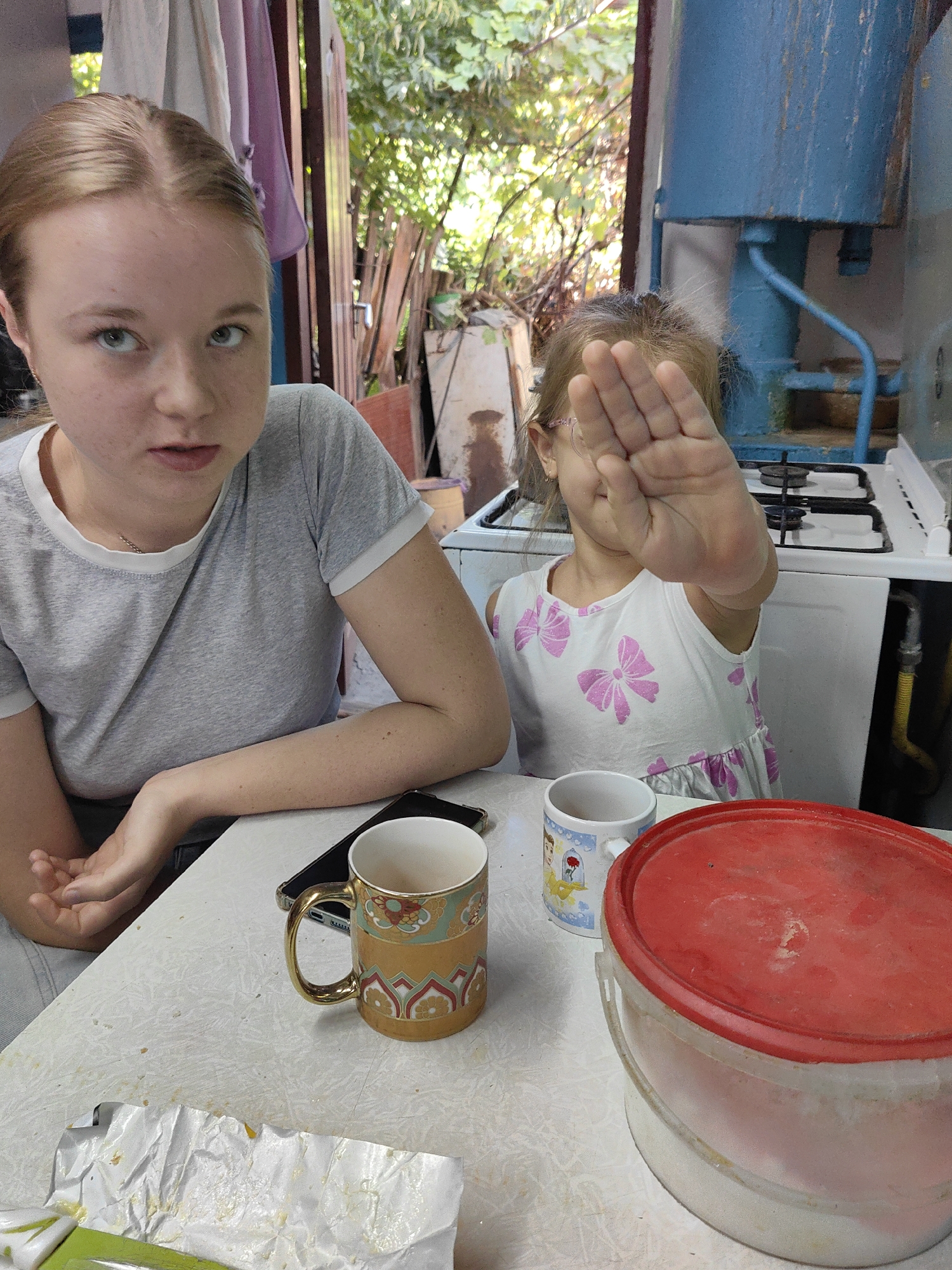 The image depicts an indoor scene, likely a kitchen or dining area, possibly in Shymkent, Kazakhstan. Two individuals are present at a table. On the left, a young woman with blonde hair pulled back is looking towards the camera with a slightly displeased expression. She is wearing a grey t-shirt. To her right, a young girl, mostly obscured by her own hand which is raised to block the camera, is wearing a white dress with pink bow patterns. She appears to be wearing glasses. On the table are several objects including a mobile phone, two mugs, and a large red-lidded container. In the background, a white gas stove is visible, along with some plumbing and greenery suggesting an outdoor or semi-outdoor setting beyond an open doorway. The environment appears somewhat rustic and worn.