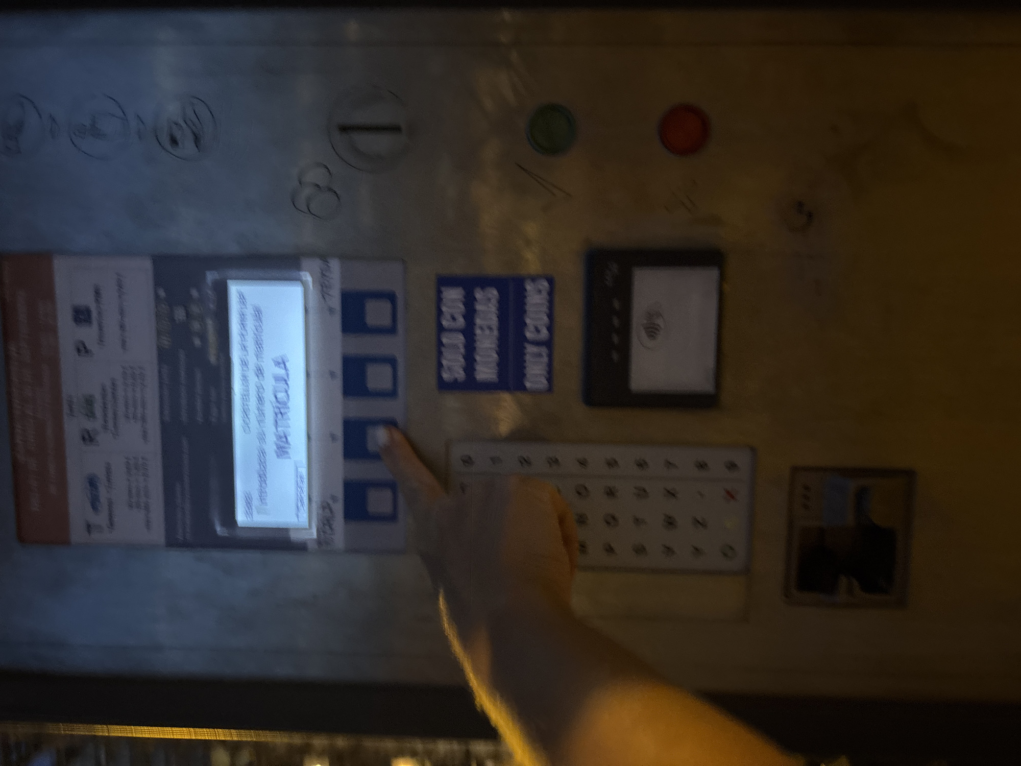 The image shows a close-up of a parking payment machine in Calp, Spain, likely outdoors. A person's hand is visible on the right side of the frame, with their index finger pressing one of the buttons on the machine. The machine has a digital display showing "MATRICULA" and "Introduzca el número de matrícula" which translates to "Enter the license plate number." There are also various buttons, slots for coins, and what appears to be a contactless payment reader. The machine itself is metal and shows signs of wear and tear, with some graffiti or markings on its surface. The background is blurred, but suggests an urban environment, possibly a street or parking lot. The lighting suggests it might be dusk or nighttime.