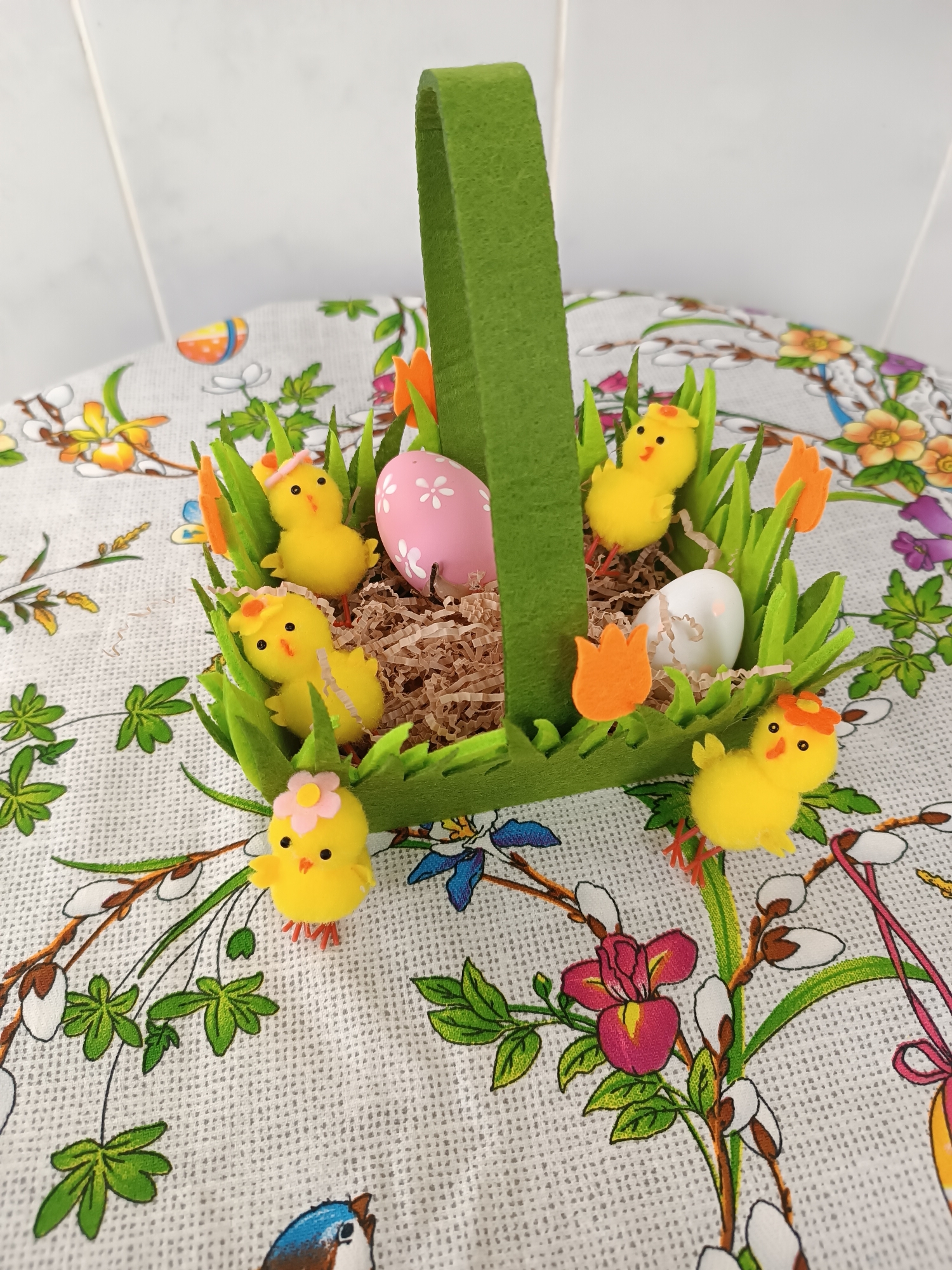 The image depicts a decorated Easter basket filled with Easter-themed decorations. The basket itself is made of green felt, with a handle extending upwards. Inside the basket, there are faux grass blades, small yellow chicks, two Easter eggs (one white, one pink with white floral patterns), and shredded brown paper resembling a nest. Some of the chicks are adorned with miniature hats or flowers. The basket sits on a tablecloth with a floral pattern featuring various flowers, leaves, and branches. The background behind the tablecloth is a plain white tiled surface. There is no location visible from the image.