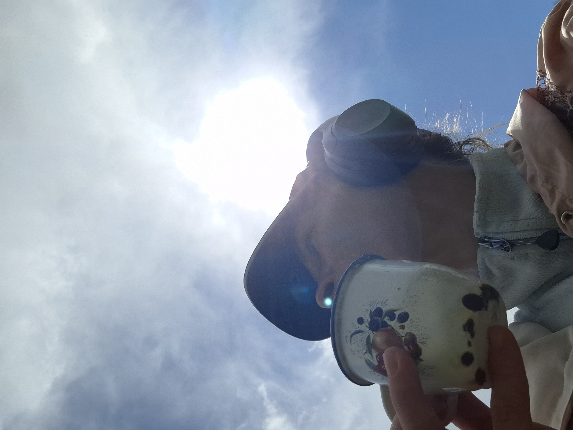 An upward-facing, low-angle photograph captures a person set against a bright, sunlit blue sky with scattered white clouds. The intense sunlight in the upper central portion of the frame creates a strong silhouette effect and significant lens flares. The person, partially visible in the upper right, wears what appears to be a wide-brimmed hat or head covering and light-colored outerwear. Dark hair is visible above the hat. They are holding an enamel mug with both hands, positioned as if drinking from it or preparing to. The mug is cream-colored and decorated with a floral or fruit pattern featuring blue berries/cherries and leaves, along with some reddish-orange elements. Several dark, irregular spots or chips are visible on the mug's surface. The bright sun suggests it is daytime, likely midday. No text is visible in the image. The scene is set in Елец-Маланино, Russia.