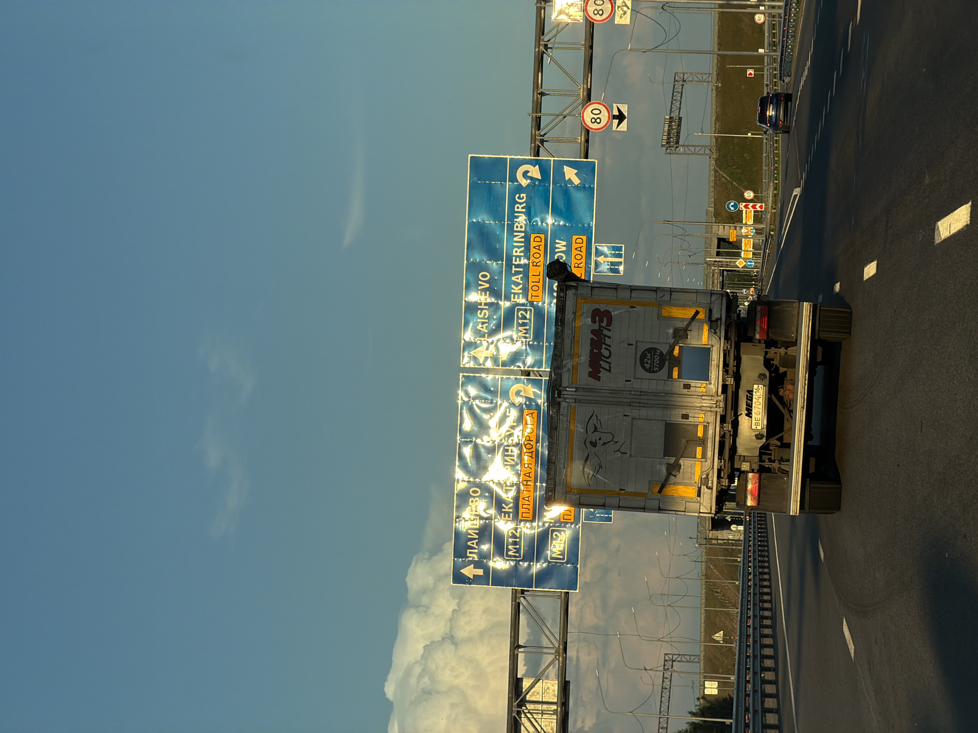 A truck travels down a highway in Egor'evo, Russia, under a clear blue sky with large, white clouds. The truck, with a graphic of a bull on its side, is positioned in the right lane, moving away from the viewer. Overhead signs indicate directions to "LAISHEVO" and "EKATERINBURG" with "TOLL ROAD" highlighted in orange. The highway has multiple lanes and is bordered by a metal guardrail and lampposts. In the distance, other vehicles, including a dark SUV, can be seen on the road. Speed limit signs showing "80" are visible on the right side of the highway. The sun appears to be setting, casting a warm light on the scene.