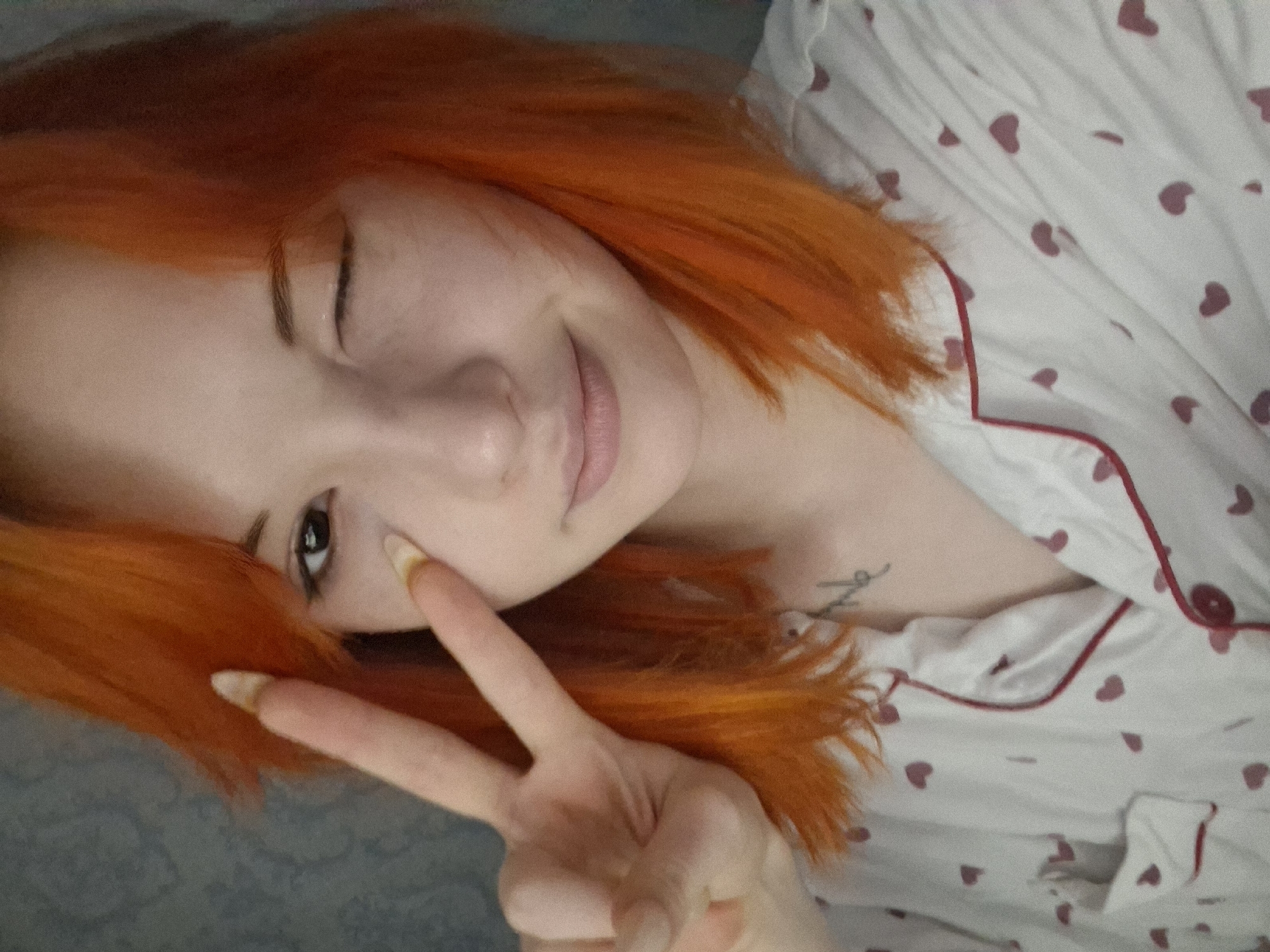 A young woman with striking bright orange-red hair poses for a selfie in an indoor setting, identified as Chaikovsky, Russia. She is lying down or reclining, indicated by the tilted perspective and the soft, textured surface visible in the background. Her fair skin contrasts with her vibrant hair.
The woman is making a playful expression, winking with her right eye and smiling subtly with closed lips. She extends her right hand towards the camera, forming a peace sign (V-sign) with her index and middle fingers. Her fingernails are long and appear to have a natural or light, possibly worn, appearance.
She is dressed in a light-colored pajama top, adorned with numerous small, dark pink heart patterns and featuring red piping along the collar and front placket. A dark pink button is also visible on her top. A small, cursive tattoo is partially visible on her left clavicle or upper chest area; the text is obscured and illegible. The overall scene conveys a relaxed and lighthearted mood.