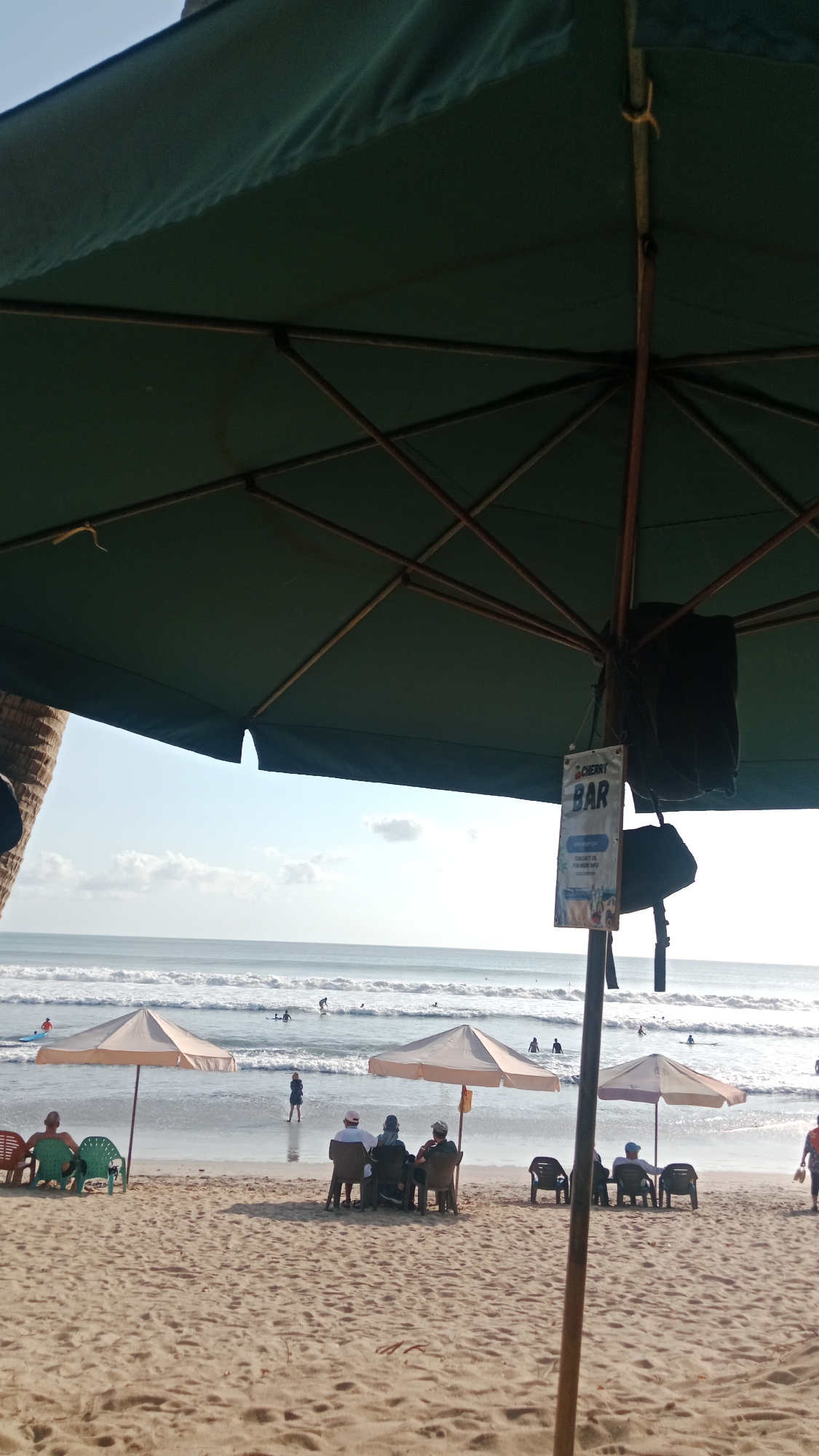 This image depicts a view from under a large, dark green beach umbrella, looking out towards the ocean. The umbrella's structure of dark poles and crossbeams is visible in the foreground. A sign hangs from the umbrella pole, reading "CHERRY BAR" with smaller text below.
On the sandy beach, several beach umbrellas are set up. To the left, two light-colored umbrellas shade two people sitting in colorful chairs. In the center, two more light-colored umbrellas are positioned behind a group of four people sitting at a table. To the right, another umbrella shades two people in lounge chairs.
The ocean in the background has moderate waves and several people are visible in the water, some appearing to be surfing. The sky is bright with a few scattered clouds. The location appears to be Kuta, Indonesia, a popular beach destination known for its surf and beachside establishments. The overall scene suggests a relaxed atmosphere at a beach bar on a sunny day.