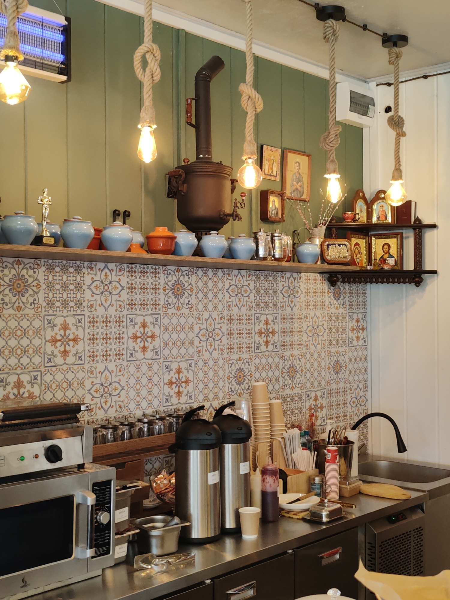 The image depicts a kitchen or cafe setting in Shikhovo, Russia. The background features a light green, paneled wall with decorative tiles below. Several hanging lightbulbs with rope accents illuminate the scene. On shelves, there are various items including blue ceramic pots, a samovar, religious icons, and decorative frames. In the foreground, a stainless steel countertop holds a microwave, a grill, two large thermoses, stacks of paper cups, and a sink with a gooseneck faucet. The overall atmosphere is domestic and slightly rustic. There are no people present in the image.
