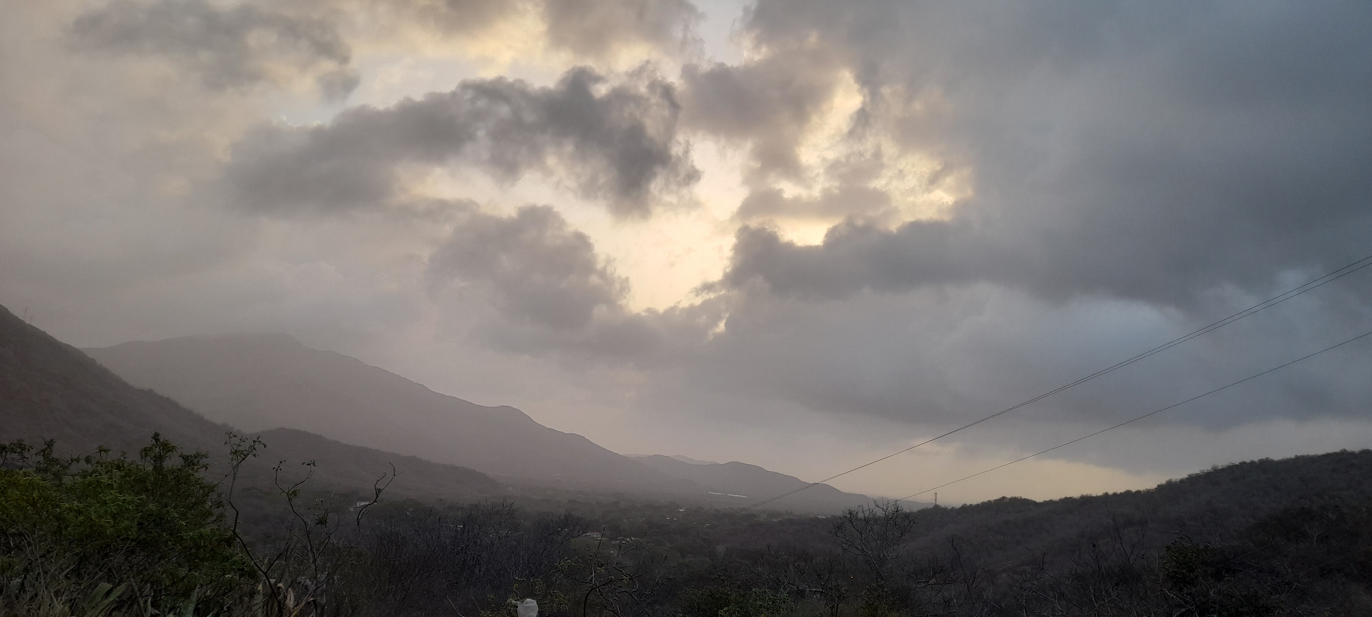 This expansive landscape photograph captures a mountainous or hilly terrain under a dramatically overcast sky. The upper portion of the frame is dominated by heavy, dark grey clouds, through which patches of softer, yellowish-white light break through, suggesting either dawn or dusk. This diffused light illuminates the distant parts of the scene.
Multiple layers of mountains and hills stretch across the image, receding into the background. The closest mountains on the left display some dark green vegetation, while successive ranges appear progressively lighter and hazier, blending into the atmosphere and appearing almost silhouetted or shrouded in mist.
In the foreground and mid-ground, the landscape is covered with sparse vegetation, including trees and shrubs, many of which appear bare or have minimal foliage, suggesting a dry season or a specific type of arid flora. A small area of greener foliage is visible on the far left foreground.
Man-made elements include three distinct power lines stretching horizontally across the middle-right section of the sky. Faint outlines of what appear to be small settlements or buildings can be discerned in the valleys between the distant hills. A utility pole or structure is barely visible on the distant right horizon, beneath the power lines.
The overall mood is quiet and vast, characterized by muted tones of grey, brown, and subtle greens, highlighted by the soft light filtering through the clouds. No visible text is present in the image.