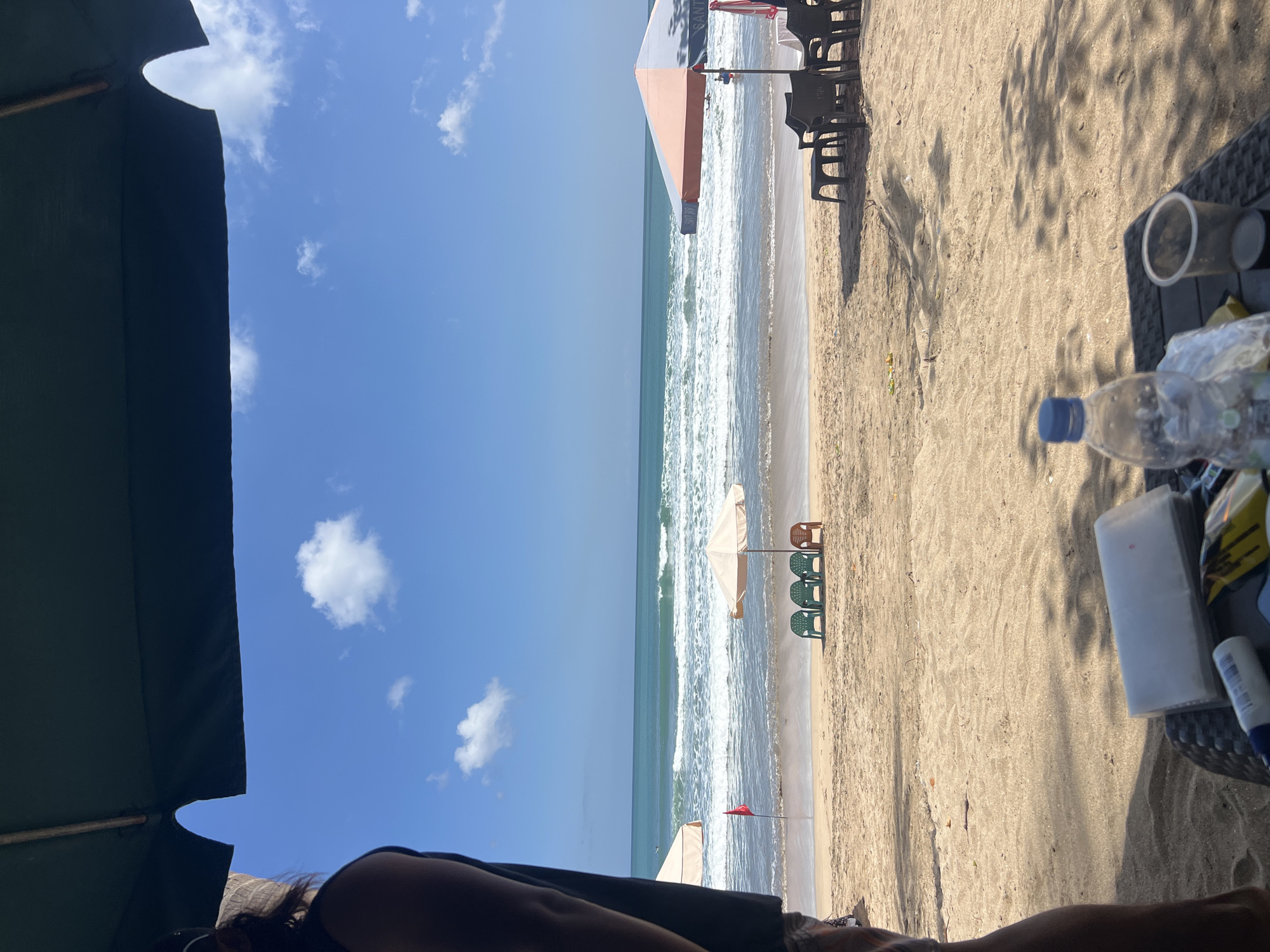 This is a beachfront view of Kuta Beach in Bali, Indonesia, on a sunny day with a clear blue sky and scattered clouds. In the foreground, a person's left arm and shoulder are visible, obscured by a large dark green umbrella, suggesting the viewer is seated under shelter. A woven table in front of the person holds a plastic water bottle with a blue cap, a clear plastic cup, a white rectangular container, and some yellow packaging. The sandy beach stretches towards the ocean, with gentle waves rolling onto the shore. Several beach chairs and umbrellas are visible on the sand. To the left, a light-colored umbrella shades a red flag. To the right, a large umbrella with orange and grey panels is set up, with dark plastic chairs and a low table nearby. In the distance, the turquoise ocean meets the horizon under the bright sky.