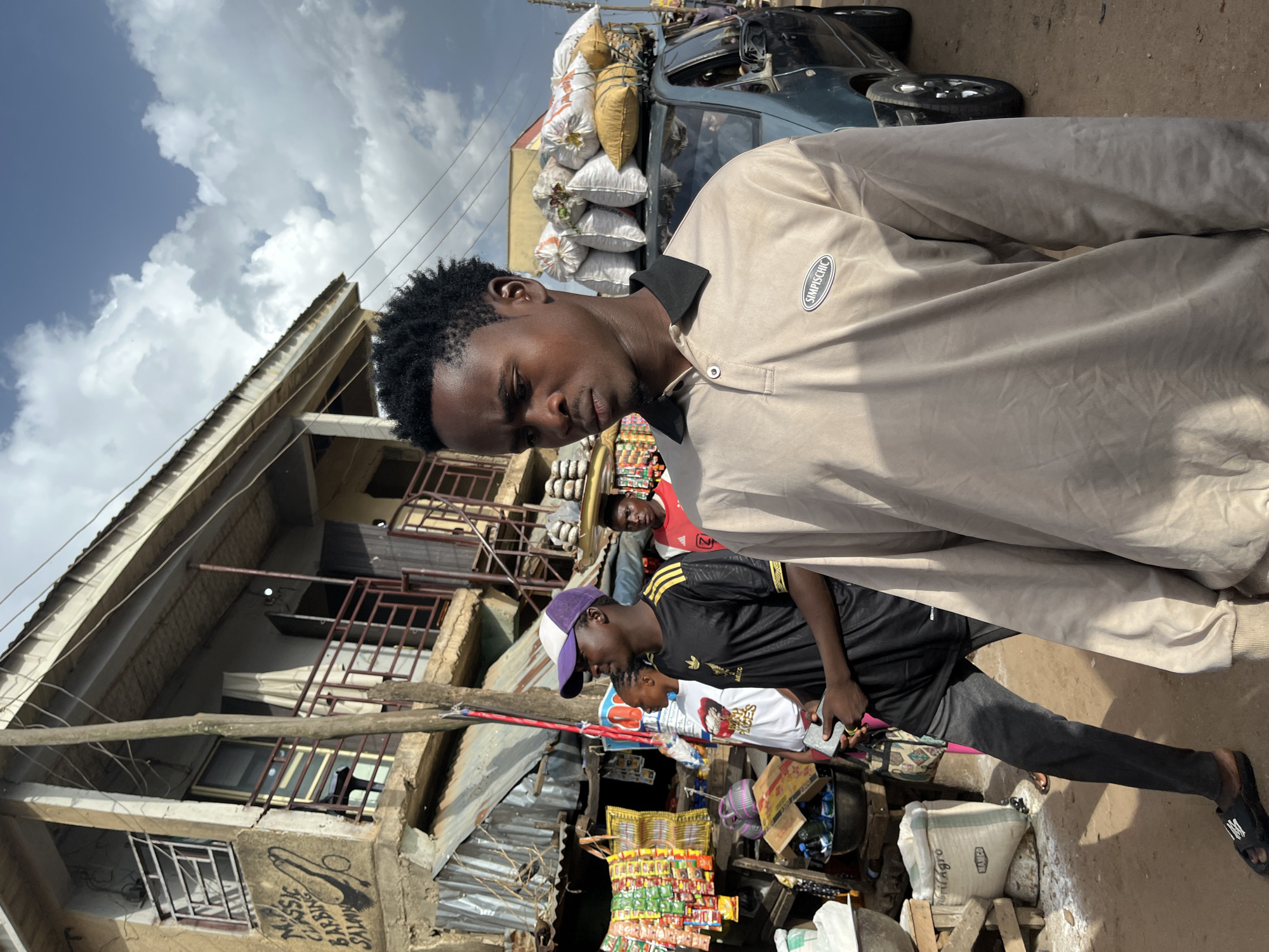 In Rafin Par, Nigeria, a bustling outdoor market scene unfolds. The foreground is dominated by a young man in a light brown polo shirt with the brand "SIMPISCHIC" on the left chest. He is looking to his right with a serious expression. Behind him, another young man wears a black and yellow striped t-shirt and a purple and white baseball cap, holding a gray object in his hands. Further back, a man in a red t-shirt is visible. In the background, a teal car is loaded with several white sacks on its roof. The market stalls display an array of colorful goods, including packaged food items, bags of produce, and various other wares. The architecture suggests a mix of older and newer structures, with some buildings showing signs of wear. The sky above is partly cloudy, with dramatic formations adding to the dynamic atmosphere of the scene. The ground appears to be unpaved, typical of a marketplace in Nigeria.