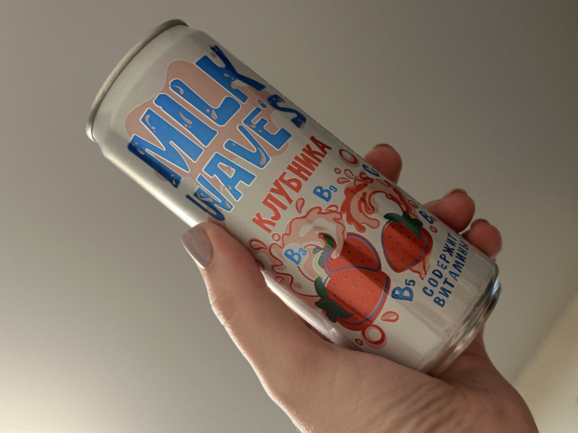 A light-skinned hand, with nails painted in a muted taupe or mauve shade, grips an aluminum beverage can vertically against a plain, light grey background. The can's label features the brand "MILK WAVES" in blue, stylized text, set on a pink, wavy background. Below this, "КЛУБНИКА" (Strawberry) is written in red Cyrillic characters. The design includes illustrations of red strawberries and white/pink milky or creamy swirls. Visible vitamin identifiers on the label include B3, B9, B5, and C, alongside the Cyrillic text "СОДЕРЖИТ ВИТАМИНЫ" (Contains Vitamins). The hand's thumb is positioned on the can's upper left side, with fingers wrapping around the lower portion. The lighting is soft and even. This scene is situated in the city of Chaikovsky, Russia.