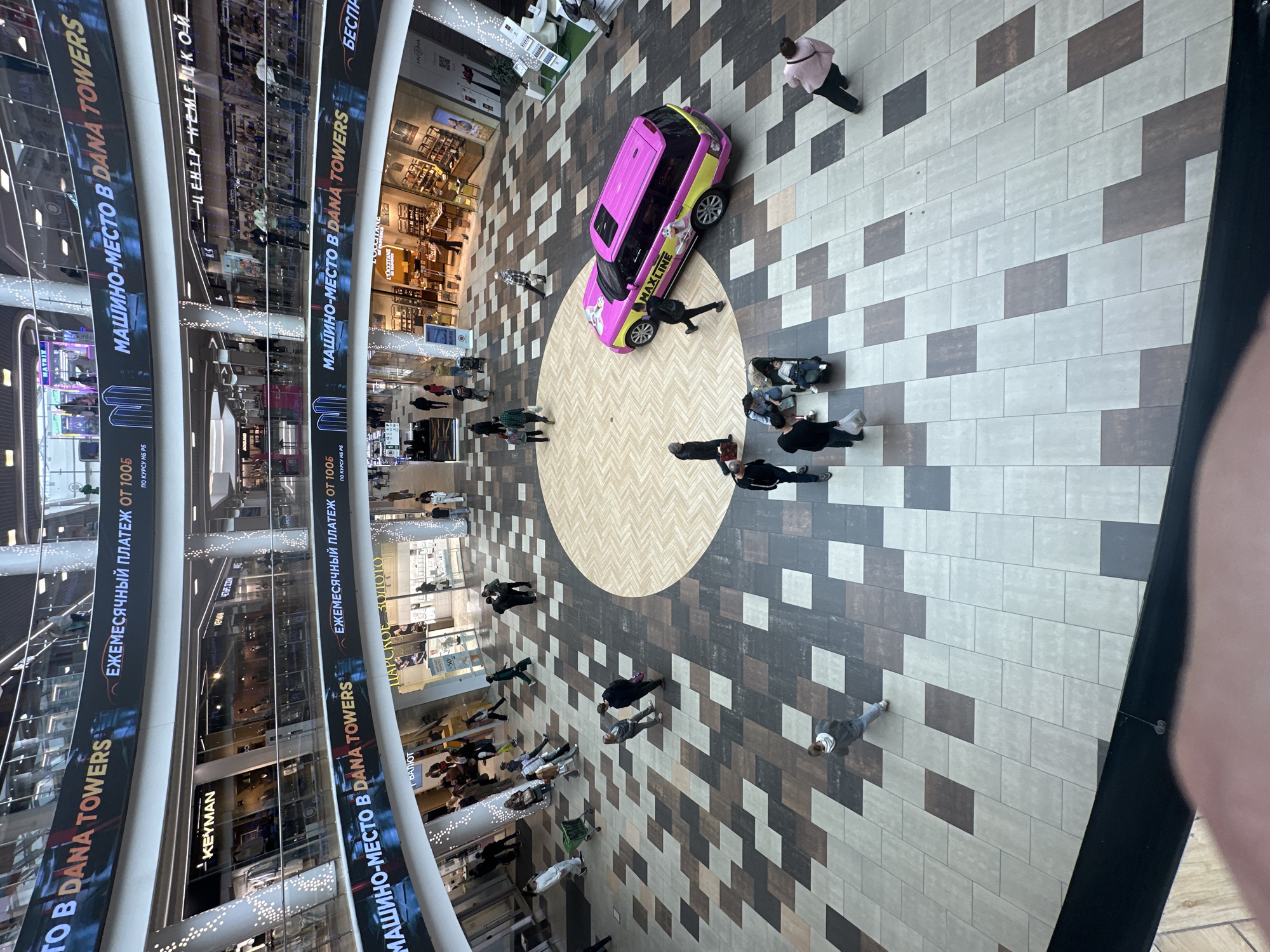 The image depicts an interior view of a multi-level shopping mall, likely in Minsk, Belarus, given the presence of Russian text on prominent signage. The scene is captured from a high vantage point, looking down into the central atrium of the mall.
The atrium floor is paved with a geometric pattern of square tiles in varying shades of brown, gray, and beige, arranged in a staggered grid. A large circular area in the center of the floor features a lighter-colored wood-grain pattern.
Dominating the lower portion of the atrium is a brightly colored, pink and yellow SUV, branded with "MAXLINE" in large letters and featuring cartoonish graphics. People are observed walking and standing around the atrium. Several individuals are seen walking on the tiled floor, some singly and others in small groups. In the background, on the upper levels of the mall, various storefronts are visible, with some displaying text in Russian, such as "KEYMAN" and "ЛАРСКОЕ ЗОЛОТО" (Larsky Gold). Banners with Russian text, including "МАШИНО-МЕСТО В DANA TOWERS" (Parking space in DANA TOWERS), are suspended across the upper levels. The mall environment appears modern, with sleek railings and a spacious design.