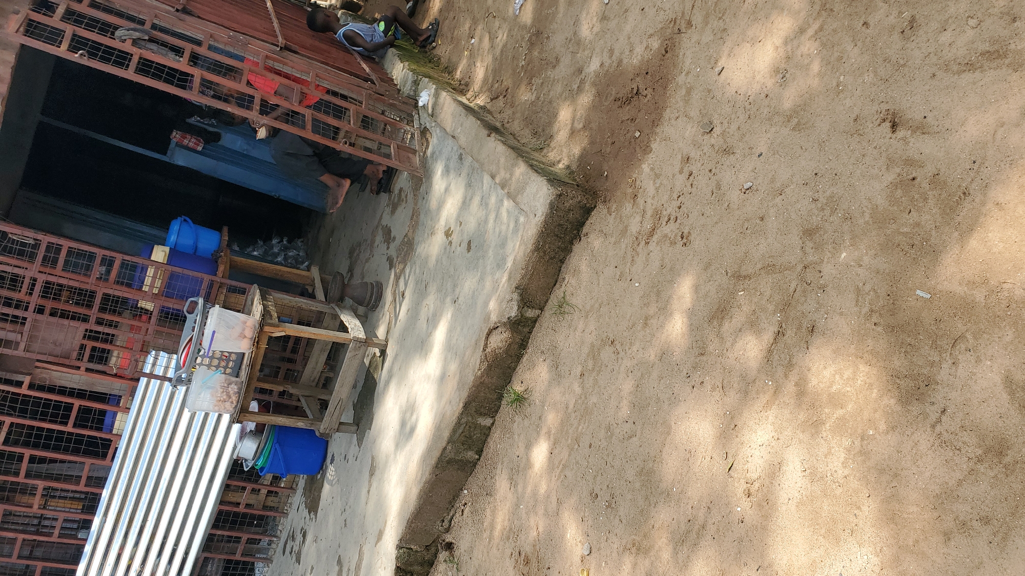 This image depicts an outdoor scene in Jalingo, Nigeria. In the foreground, sandy ground with scattered debris is visible. A raised concrete platform runs horizontally across the middle ground, with the foreground sandy area meeting it. Behind the platform, a wooden table with two clear plastic containers filled with food items sits. To the left of the table, two blue buckets and a pot are stacked. A corrugated metal sheeting is leaning against a metal grate structure behind the table. In the background, a shop entrance is partially visible, with a person sitting just inside, appearing to eat. To the right of the shop entrance, a young boy in a blue tank top and shorts is sitting. The overall environment suggests an informal market or roadside stall setting.