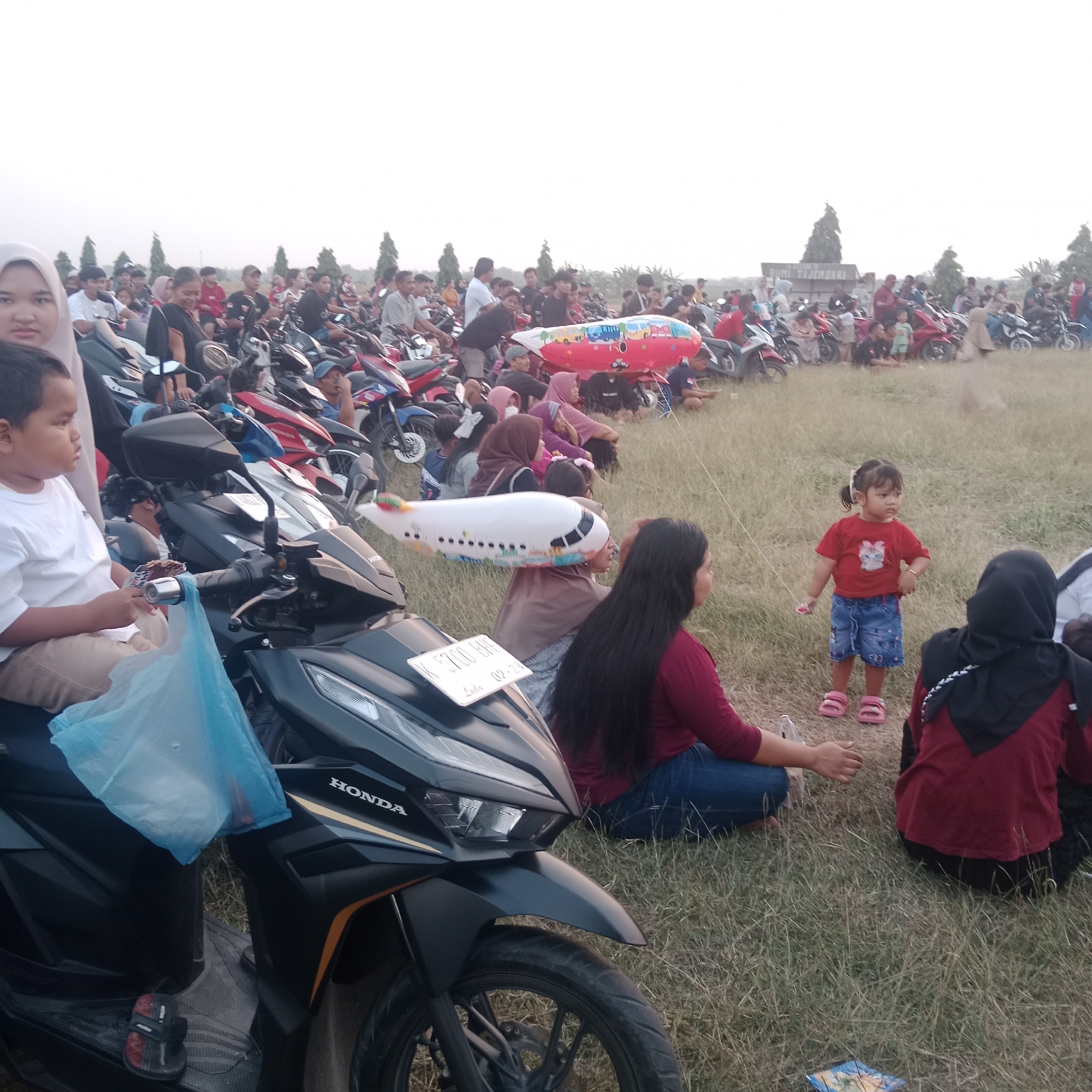This image depicts an outdoor gathering in Tegowanu Wetan, Indonesia, likely a community event or festival. A large number of motorcycles are parked in the background, suggesting a popular event that many people have arrived at by vehicle. The foreground features a group of people, including children and adults, sitting on the grass. Some individuals are holding or interacting with inflatable toys, including a large red airplane-shaped balloon and a smaller white airplane balloon. A young child in a red shirt and blue shorts is standing and looking to the side. Another child in a white shirt and light-colored pants is sitting on a motorcycle and holding a treat. The overall atmosphere appears casual and festive, with people enjoying a warm day. The environment is grassy with some trees visible in the distance. A sign that says "BUMI TAJENSARI" is partially visible in the background. The lighting suggests it is either early morning or late afternoon.