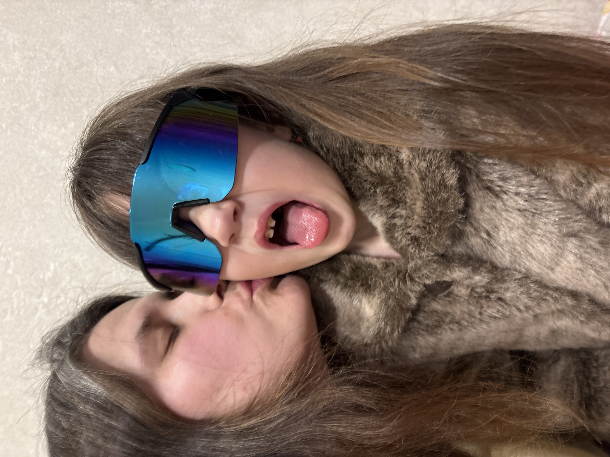 Two females are depicted in a close-up indoor shot, originating from Chaikovsky, Russia. On the left, an older female with long brown hair and closed eyes is shown in profile, puckering her lips to kiss the right cheek of a younger female. The younger female wears oversized, iridescent blue-green reflective sunglasses that obscure her eyes, and a brown fur-textured coat. Her mouth is open with her tongue extended, revealing upper primary teeth with some discoloration. The background consists of a light-colored, subtly textured wall.