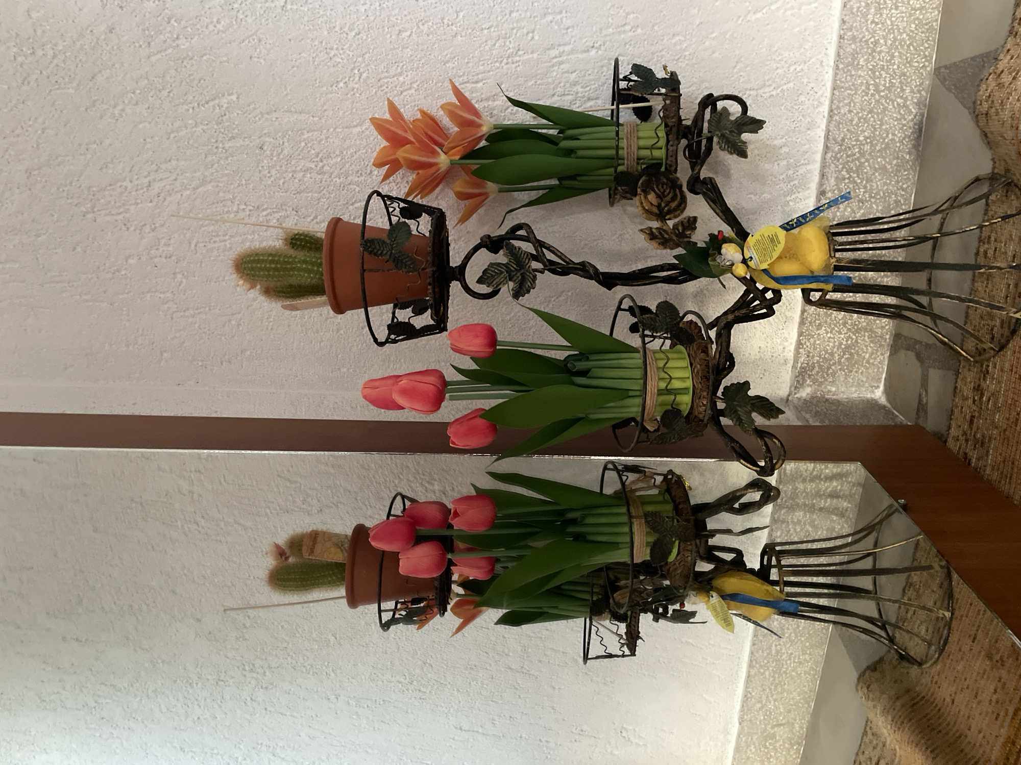 The image shows a decorative metal plant stand with multiple tiers holding potted plants and ornamental figures. On the top tier, there are two small potted cacti. The middle tiers hold potted tulips, with red tulips on the left and orange tulips on the right. The lower part of the stand features decorative metalwork and two yellow bird-shaped ornaments. The plant stand is placed in front of a textured white wall, and a mirror reflects the scene to the left. A brown carpet is visible in the foreground. The scene appears to be an interior setting, possibly a home or a shop. The presence of a mirror suggests a reflective surface is intentionally included in the display.