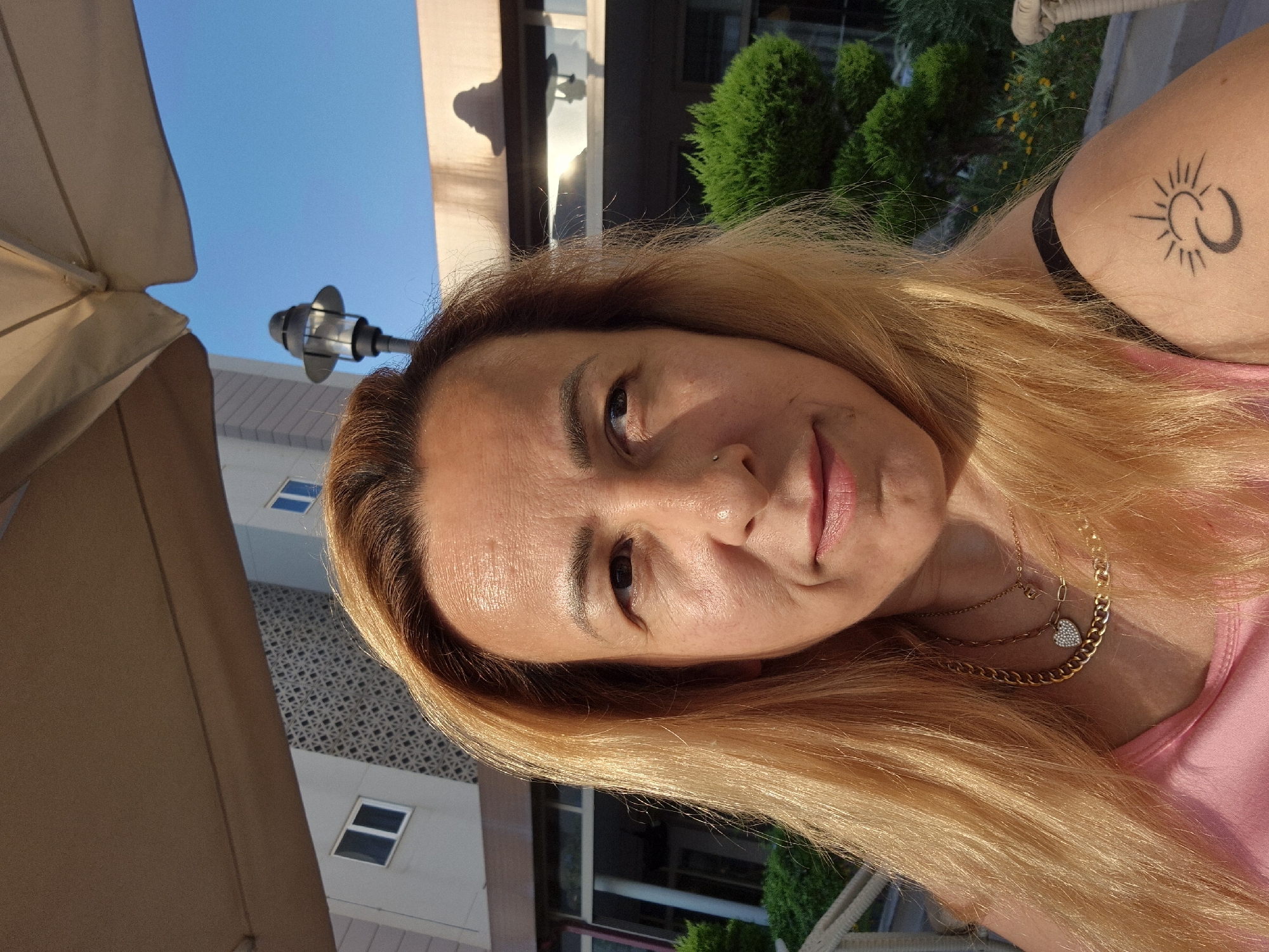 A medium shot shows a fair-skinned person with blonde hair pulled back from their face, wearing a pink tank top and gold necklaces. The person's right shoulder is visible, displaying a tattoo of a crescent moon and a sun. They are outdoors, with a building and a light fixture in the background. The lighting suggests it is daytime, possibly late afternoon given the warm tones.