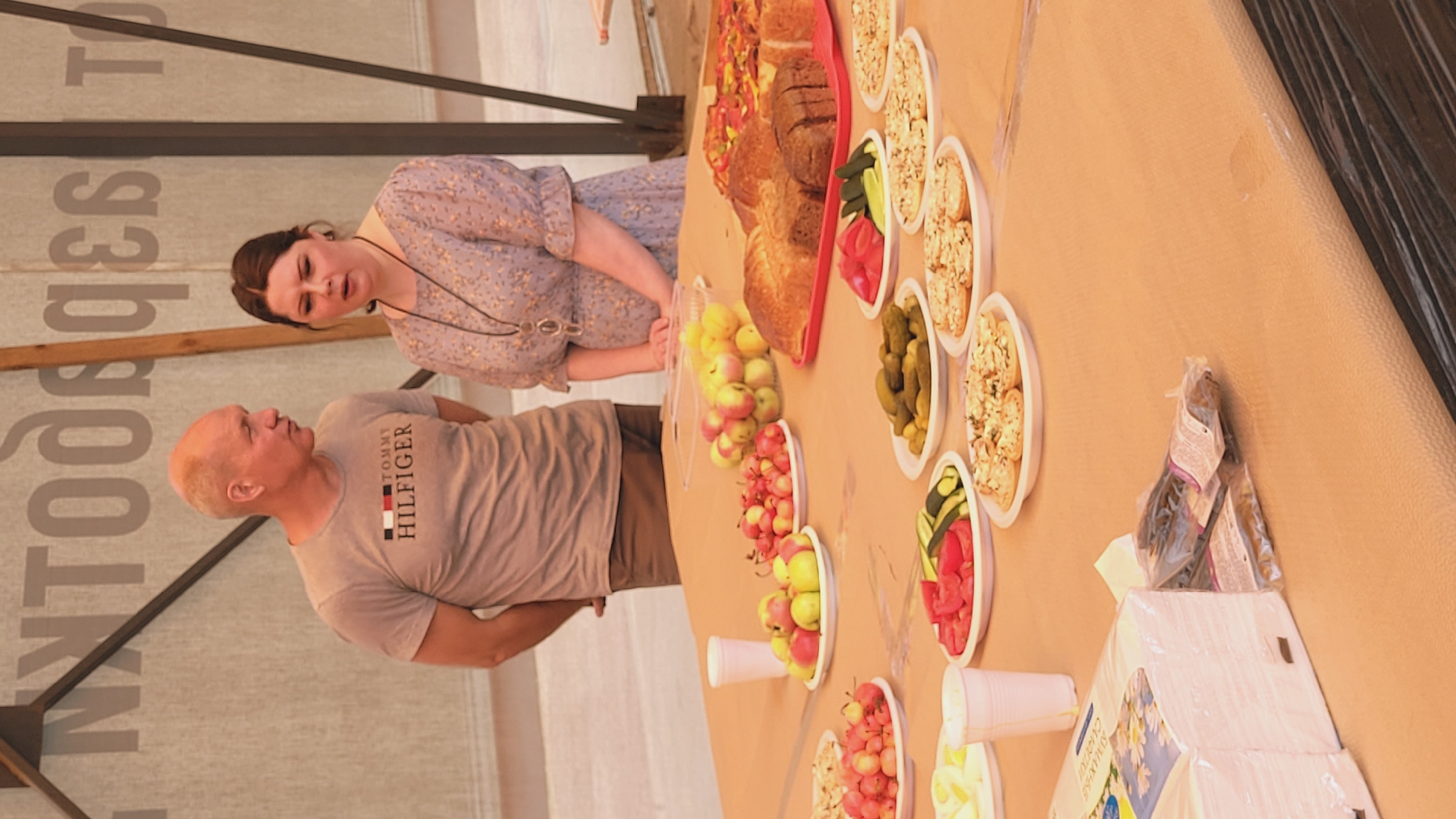 This media file depicts an outdoor gathering with a table laden with food. Two people are visible: a man with a shaved head wearing a grey t-shirt with "TOMMY HILFIGER" printed on it, and a woman with dark hair wearing a floral-patterned dress. They appear to be conversing.
The table holds a variety of food items arranged in white, disposable containers. These include what appear to be bread, sliced vegetables like tomatoes and cucumbers, pickles, and small fruits, possibly apples and berries. There are also stacks of white paper cups and some packaged goods on the table. The setting seems to be outdoors, possibly under a canopy or tent structure, with a large banner featuring text in a Cyrillic alphabet visible in the background. The overall impression is of a casual, perhaps picnic-like, event. The lighting suggests it is daytime.