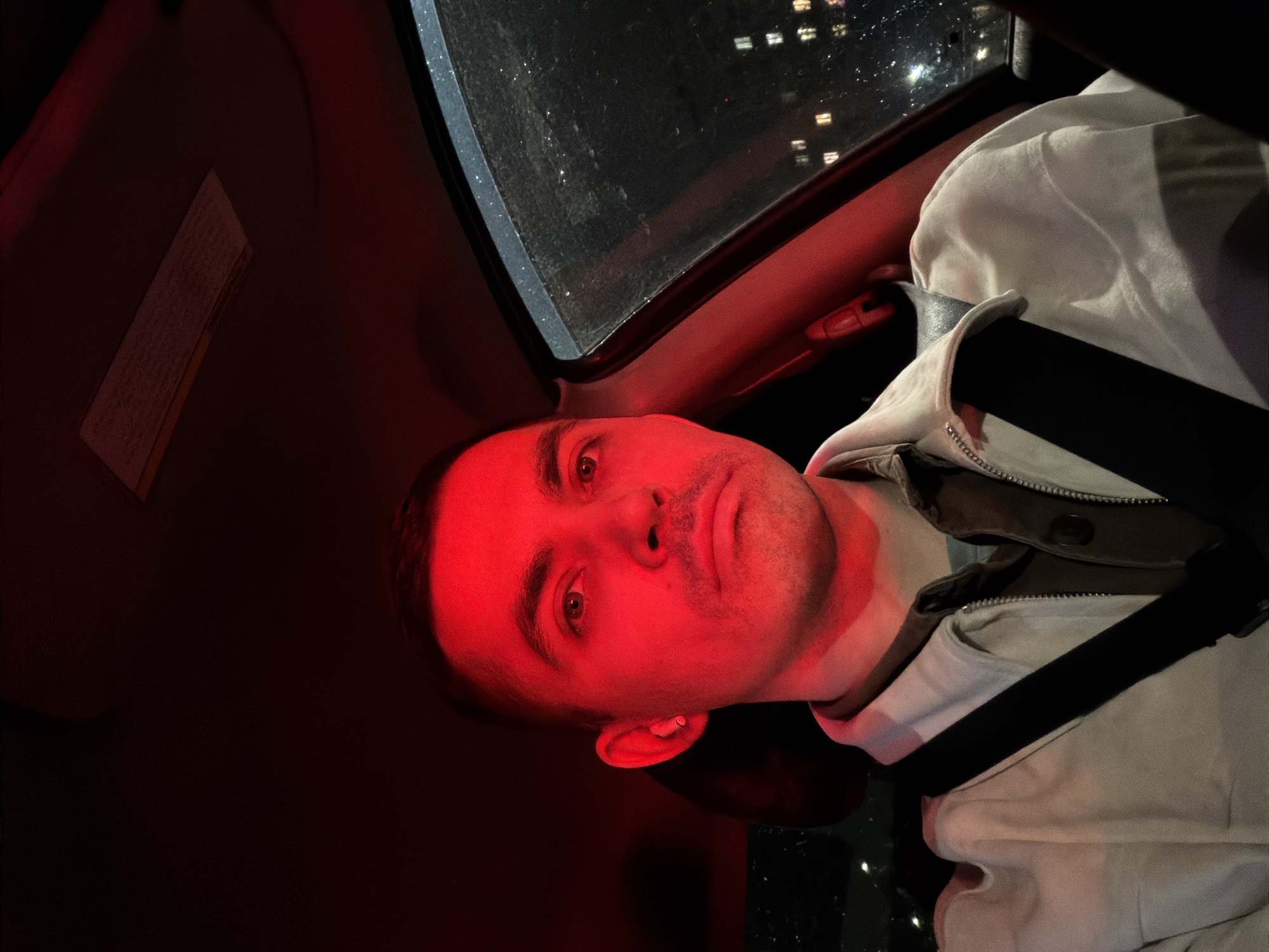 A man is sitting in a car. He is wearing a light-colored jacket and a seatbelt. The interior of the car is dark, but the man's face is illuminated by a red light. Outside the car window, there are lights from buildings, possibly indicating an urban environment. Given the red lighting and the positioning of the subject, it is reasonable to assume he is at a stop light. There is no information to confirm the location to be Izhevsk, Russia.