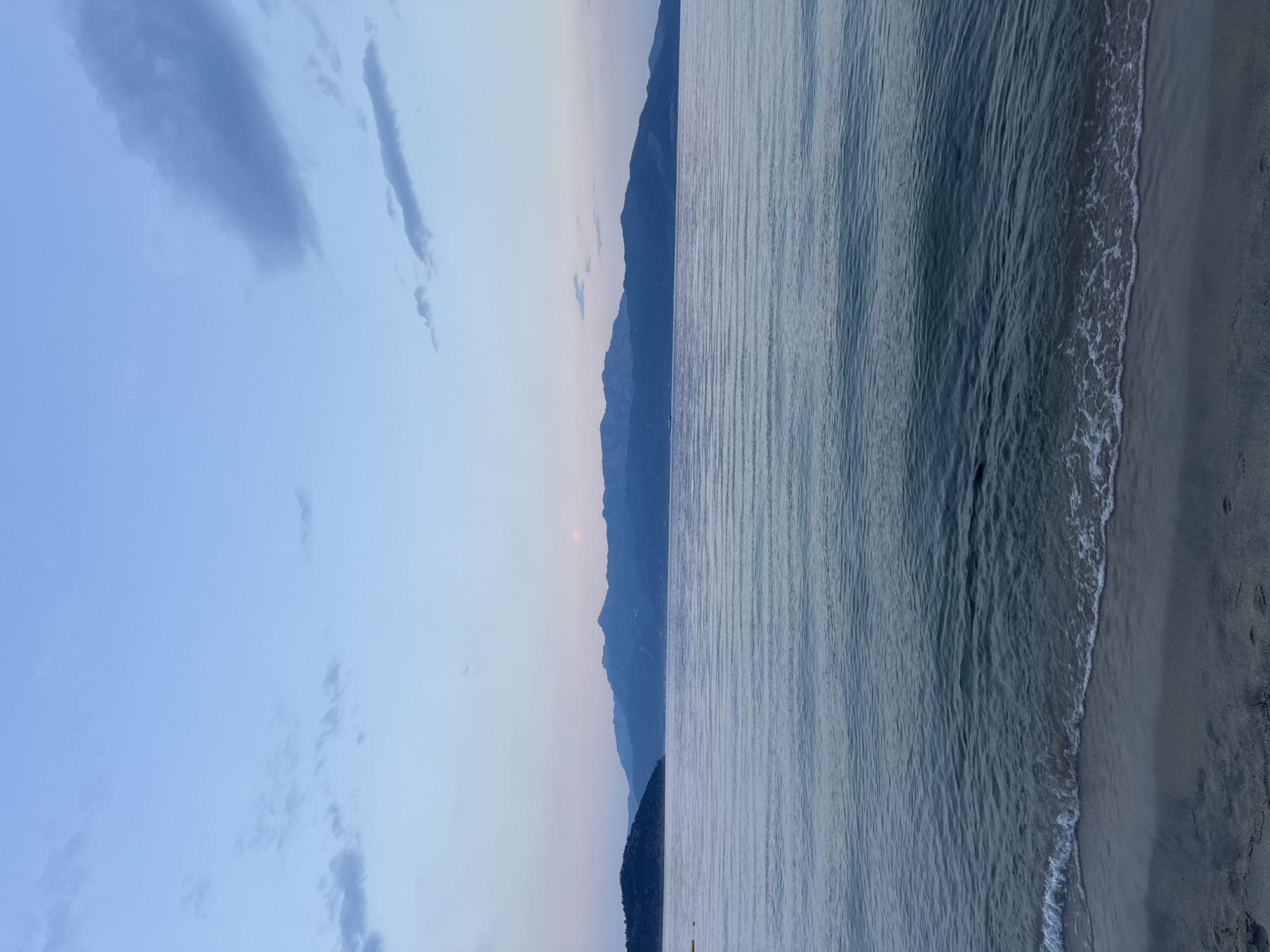 An outdoor, eye-level, full shot captures a tranquil seascape at dusk in Keramoti, Greece. The foreground features a sandy beach with gentle waves lapping the shore, their foam creating a delicate white line. The sea stretches out to the horizon, its surface rippled by the breeze. In the middle ground, a long range of dark blue mountains forms the backdrop, their silhouettes softened by the twilight. The sky above is a gradient of pale blue, with a few wispy clouds adding texture. The overall atmosphere is serene and peaceful, conveying the calm beauty of the coastline at the end of the day. There are no people or animals visible in the frame, nor any man-made structures besides what is implied by the distant mountains.