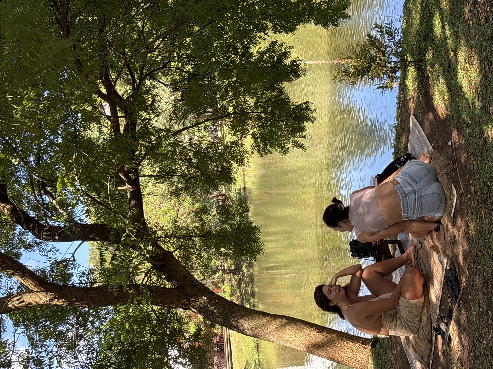 Two women are relaxing by the water's edge under the shade of a large, leafy tree on a bright, sunny day. The scene is set in a natural, outdoor environment, possibly a park or riverside area, with lush green foliage and calm water.
In the foreground, one woman with dark hair is seated on a light-colored woven mat, leaning back with her hand near her face, seemingly in thought or casual conversation. She is wearing a light-colored, possibly crocheted or knit, sleeveless outfit. Her bare feet are visible on the mat next to a pair of dark flip-flops or sandals.
Further back, closer to the water, the second woman is kneeling or sitting on a wooden structure, also on a mat. She has dark hair tied up and is wearing light-wash denim shorts and a light-colored, possibly pink or beige, short-sleeved top. She is facing towards the water, with a dark bag placed behind her. Her bare feet are visible, one resting on the mat.
The body of water, likely a lake or pond, has a greenish-brown hue and reflects the sky and surrounding trees, with gentle ripples disturbing the surface. The ground where they are seated is a mix of dirt and grass, with strong shadows cast by the large tree overhead, indicating bright daylight. The overall atmosphere is one of leisure and tranquility. No discernible text is clearly visible on any objects or clothing.
