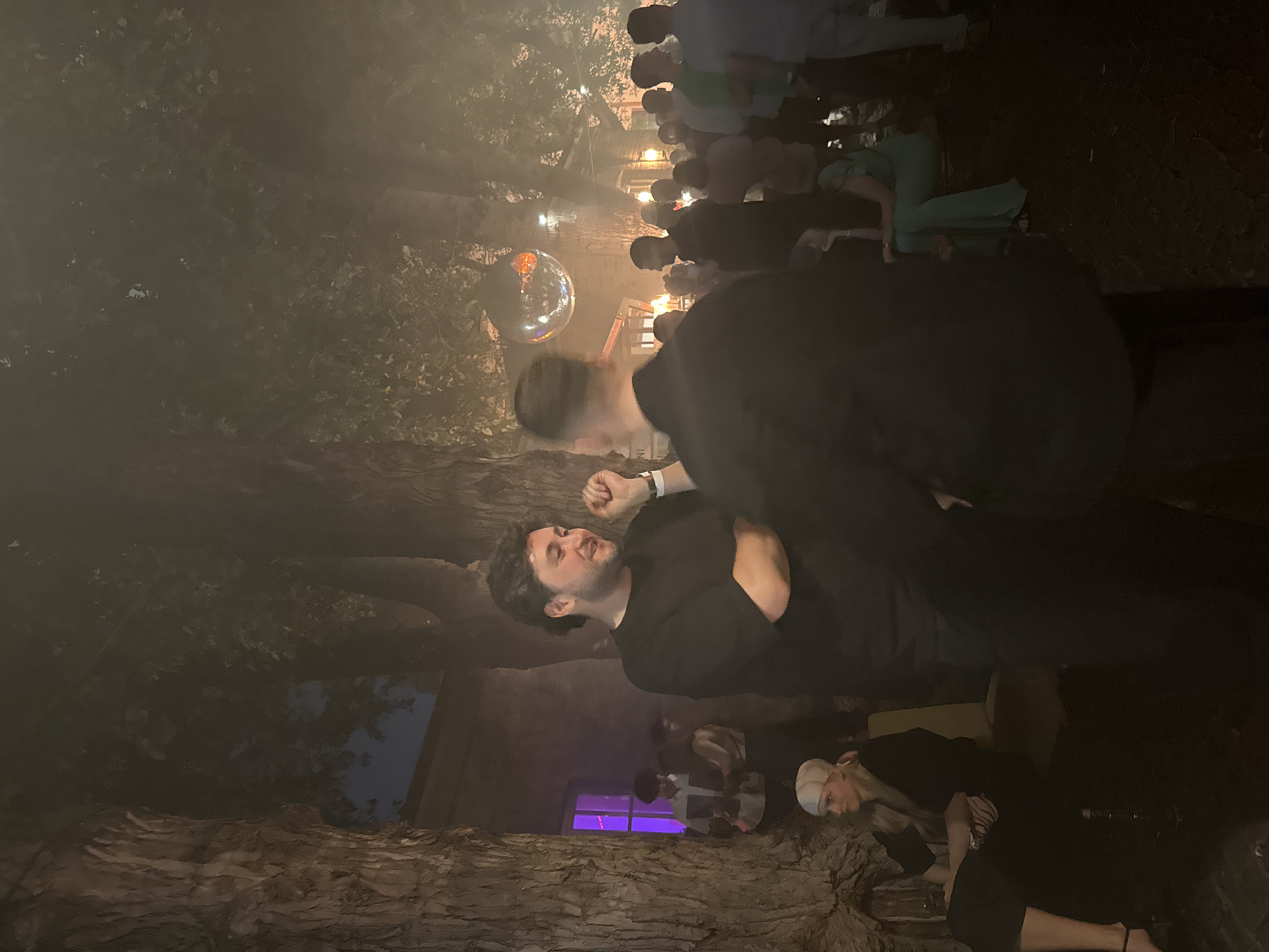 The image depicts an outdoor evening gathering in Moscow, Russia. A group of people are socializing amongst large trees with textured bark. In the foreground, two men are engaged in conversation; one man, wearing a black t-shirt, has his arms crossed and is looking towards the other man, who is facing away from the camera. Behind them, a disco ball hangs in the air, reflecting light. In the background, a crowd of people are visible, with some standing and others seated. The environment is dimly lit, with some areas illuminated by artificial lights, creating a hazy atmosphere, possibly due to fog or smoke.