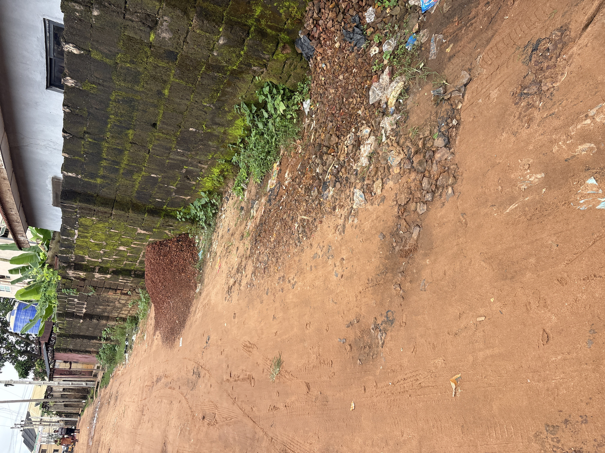 The image depicts a reddish-brown dirt road in Nkpor, Nigeria, with a large, moss-covered brick wall running along the right side. The wall appears old and weathered. Piles of reddish-brown gravel are present near the wall, and scattered debris, including plastic bags and wrappers, litters the roadside. Tire tracks are visible on the road, indicating vehicle traffic. In the background, buildings and lush green vegetation, including banana trees, are visible. Two individuals are walking away from the camera on the left side of the road, dressed in casual attire. The sky is overcast, suggesting a cloudy day.