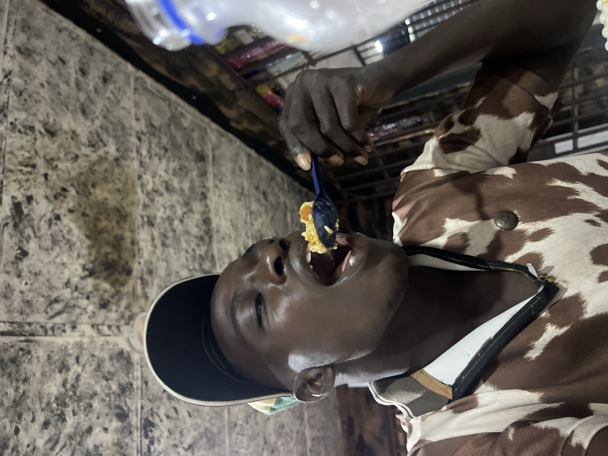 A close-up, vertically oriented shot shows a person, presumably male, with dark skin, wearing a cap and a brown and white cow-print shirt. The person is in the process of eating, with a blue plastic spoon holding a spoonful of food, which is being brought towards their open mouth. Their eyes are partially closed, suggesting focus on the food or a moment of enjoyment. The background appears to be an indoor setting with textured, possibly tiled, walls and some shelves or structures visible, hinting at a market or food stall environment. A blurry plastic bottle is also visible at the top. The lighting suggests it could be daytime, possibly indoors with artificial light. The image is sharp on the person's face and the food, with the background out of focus.