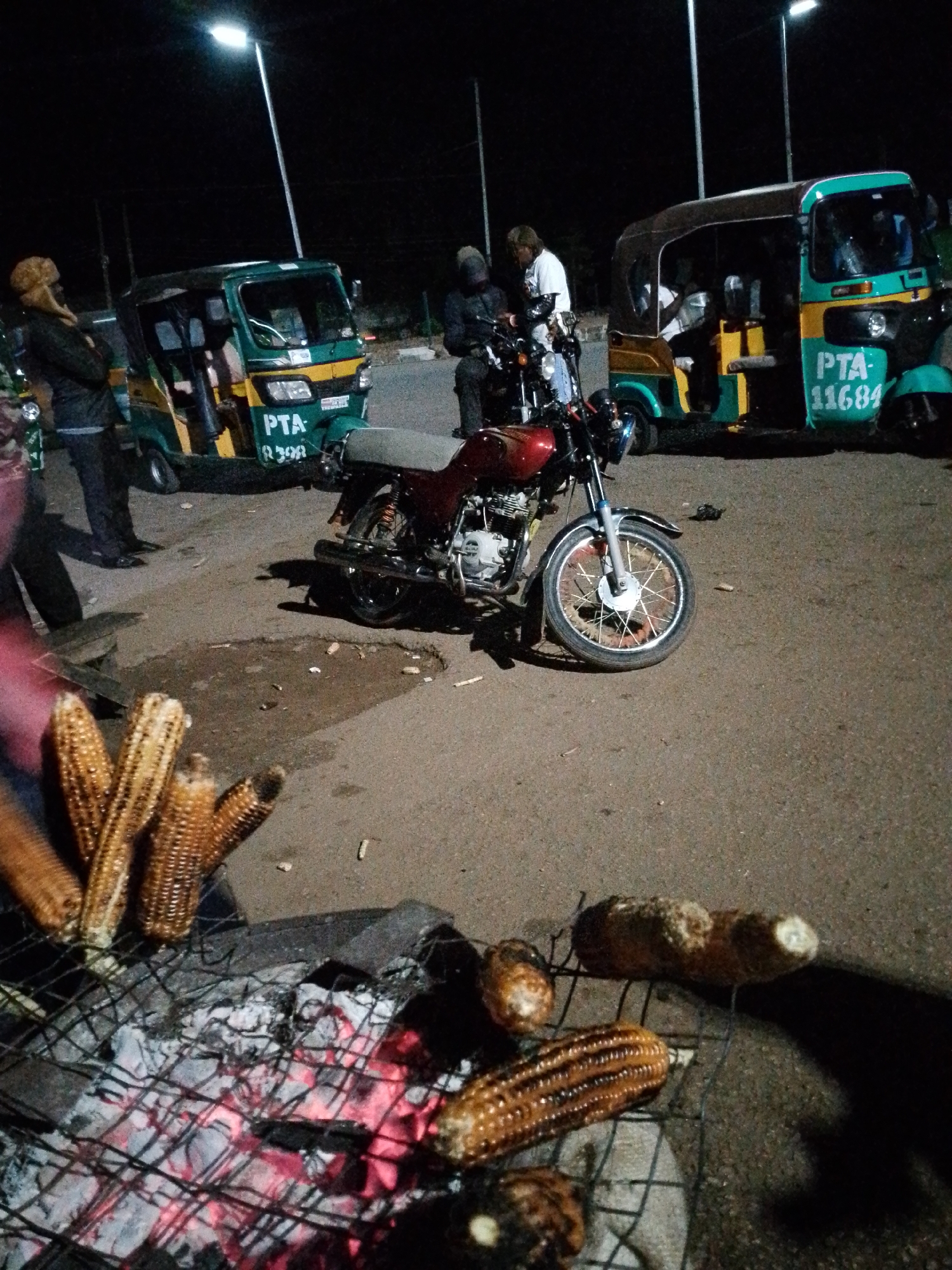 The image depicts an outdoor scene at night in Bukuru, Nigeria. The primary subjects are several tuk-tuks (auto-rickshaws) and a motorcycle, suggesting a street or roadside setting, possibly a marketplace or transportation hub. In the foreground, a charcoal grill is set up with corn being roasted. The glowing embers and the grilled corn indicate that food is being prepared and likely sold.
There are a few people visible. One person in the left tuk-tuk appears to be a passenger. Another person, wearing a white shirt and a cap, is standing near the motorcycle. A third person, partially obscured, is in the background. The overall mood appears to be a typical evening activity, with people gathered around for food and transportation.
Visible text includes license plates on the tuk-tuks. The green and yellow tuk-tuk on the left has "PTA 8398" visible. The teal and yellow tuk-tuk on the right has "PTA 11684" displayed prominently. The lighting is provided by streetlights and possibly ambient light from nearby sources, casting shadows and illuminating the scene. The presence of roasted corn suggests a local street food vendor operating in the evening. The ground is paved asphalt, with some debris scattered around. The time of day is clearly night, indicated by the dark sky and artificial lighting.