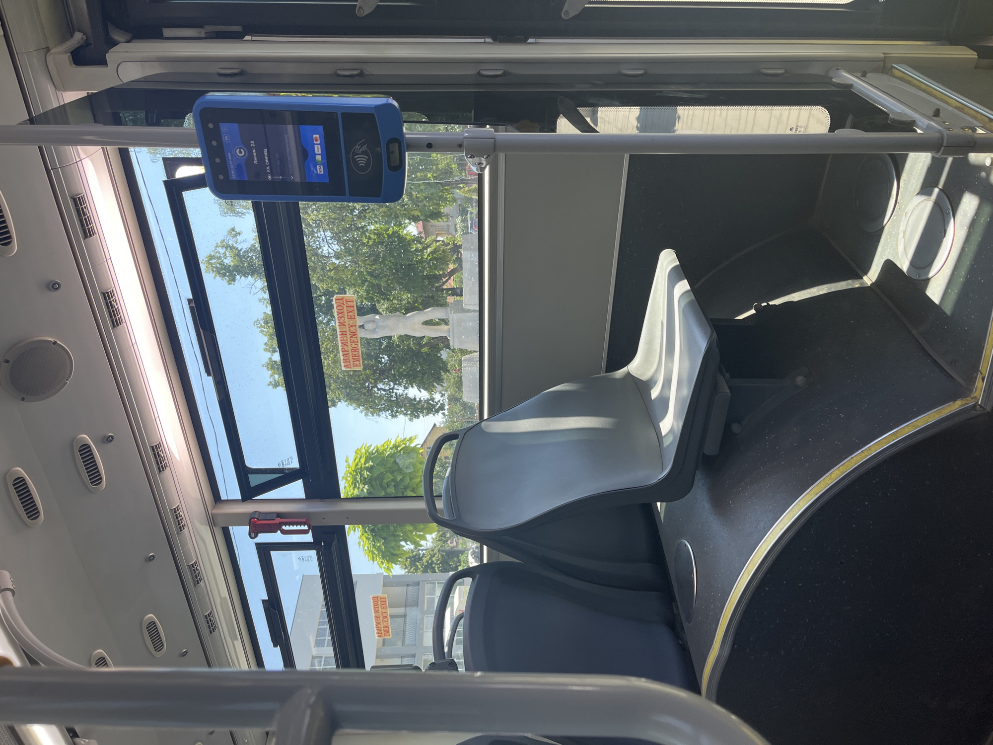 The image, taken inside a public bus in Kubratovo, Bulgaria, depicts the interior from a seated perspective, looking towards the front. Two rows of dark grey bus seats are visible. A blue ticket validation machine is mounted on a pole near the window, displaying route information and payment options. Outside the window, trees and a statue are visible, suggesting an urban setting. A red "EMERGENCY EXIT" sign is also present on the window. The bus appears to be stationary, with natural light streaming in through the windows.