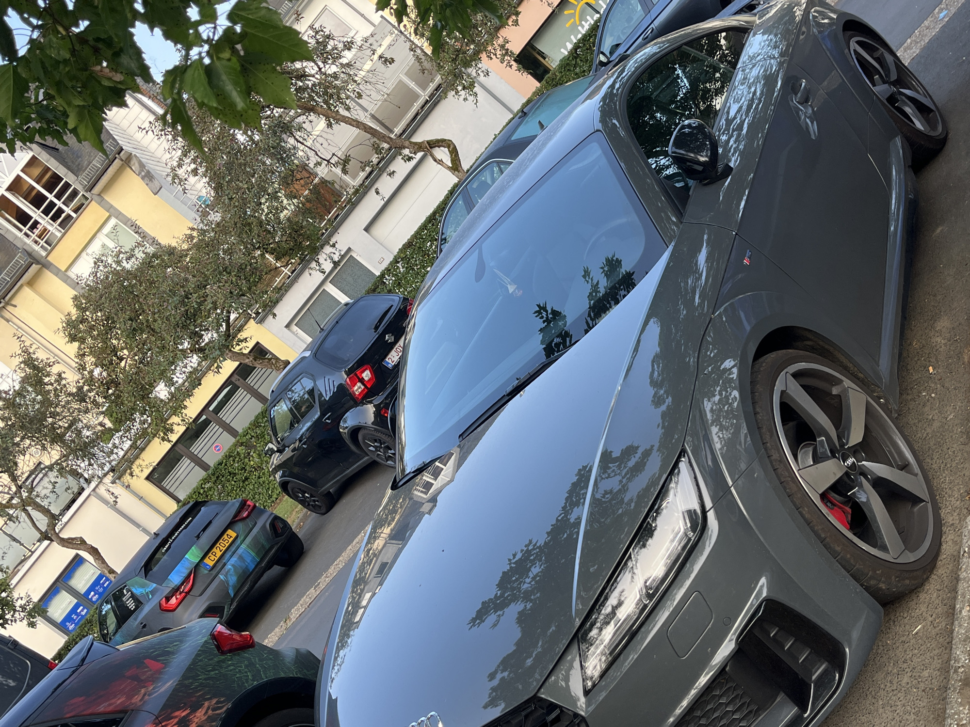 The image shows a street scene in the city of Saint-Vith, Belgium, with multiple cars parked. In the foreground, a dark grey Audi TT is parked, its hood and front quarter panel are visible. To the left of the Audi TT, a dark SUV with a colorful graphic wrap is parked. Behind the Audi TT, a black SUV or hatchback with a license plate that reads "CP 2054" is parked. Further behind that, another black vehicle is partially visible. The background consists of a building with multiple windows and trees lining the street. The overall lighting suggests it is daytime.