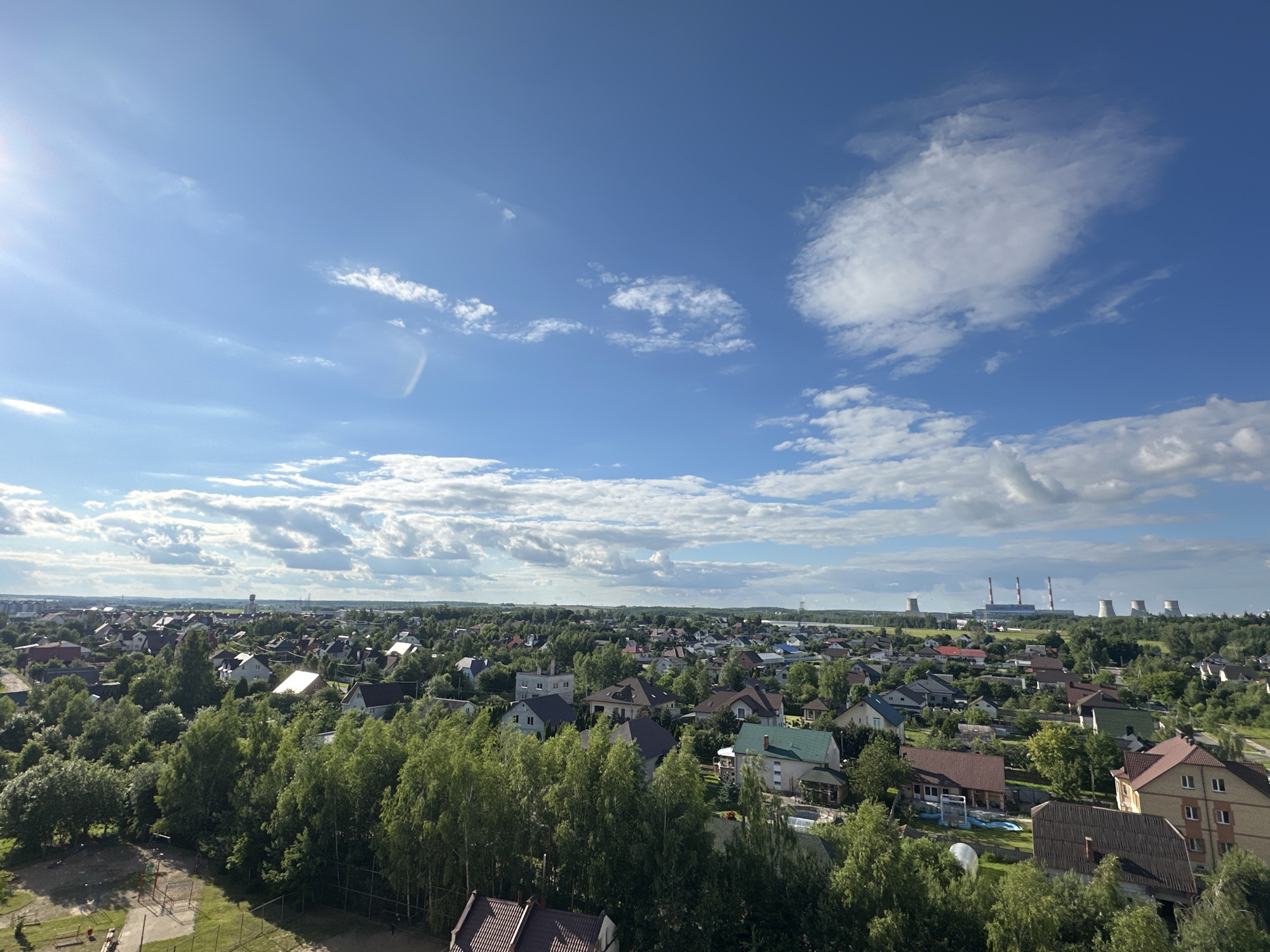 This is a high-angle, panoramic photograph of a suburban landscape under a bright, partly cloudy sky. The scene depicts a residential area filled with numerous houses, characterized by their varied architectural styles and roof colors, interspersed with lush green trees and vegetation. In the foreground, a playground with swings and climbing structures is visible, along with some low-lying structures and fencing.
The midground and background are dominated by the dense canopy of trees that surrounds and weaves through the neighborhood. Beyond the residential area, a more distant view reveals industrial structures, including power plant cooling towers and smokestacks, under the expansive blue sky. The sun is bright, suggesting it is daytime, likely late morning or afternoon, given the shadows cast. The clouds are scattered, indicating fair weather with some atmospheric texture. The overall impression is one of a developed rural or semi-urban setting in Belarus. No people are clearly discernible in the frame, and there is no visible text.