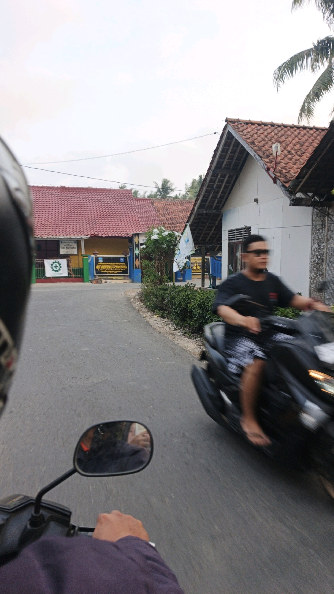 The image captures a scene from the perspective of a person riding a motorcycle, looking forward and slightly to the left. In the foreground, the hand and arm of the rider are visible, wearing a purple long-sleeved shirt, gripping the handlebars. To the left, a portion of a black helmet is in view. A rearview mirror attached to the motorcycle shows a reflection of the rider's face and the road behind.
On the right side of the road, a man is riding a black motorcycle, also in motion, with his body blurred due to the speed. He is wearing a black t-shirt and patterned shorts, and sunglasses. The motorcycle has its headlight on.
The road appears to be asphalt and is surrounded by vegetation on the right. On the left side of the road, there is a building with a red tiled roof. A sign with green and white graphics, including a gear symbol, is visible on a fence in front of the building, which appears to be a school with yellow and blue accents. Another sign indicates "SD NEGERI 4 CIRENDA," identifying the location as Cibenda State Elementary School 4. Further down the road, more buildings with red tiled roofs and white walls are visible, characteristic of a rural or suburban area in Indonesia. Palm trees are also present in the background. The sky is overcast with a light cloud cover.