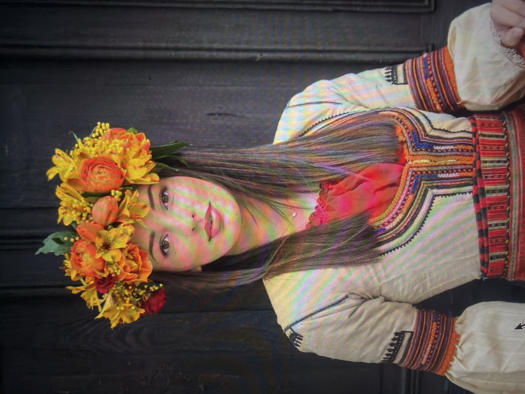 A young woman is depicted in a front-facing, waist-up portrait against a dark, unadorned wooden background, a scene set in Shumen, Bulgaria. She has long, straight dark hair and a fair complexion, with her eyes open and lips exhibiting a faint smile. Her head is adorned with a vibrant floral crown composed of numerous yellow, orange, and red flowers, interspersed with green leaves. She wears an off-white long-sleeved garment, possibly a traditional blouse, featuring intricate multi-colored embroidery. The chest and neckline display geometric patterns in hues of red, orange, blue, black, and white. The sleeves are voluminous at the wrist, with distinct embroidered bands in shades of orange, red, purple, and black above the cuffs. A segment of a red garment or lining is visible beneath the primary top at her chest. A woven, striped sash or belt, predominantly red with other colors, encircles her waist. A thin necklace with a small, singular bead or pendant is visible on her neck. The entire image exhibits horizontal color banding, indicative of a digital capture from a display screen.
