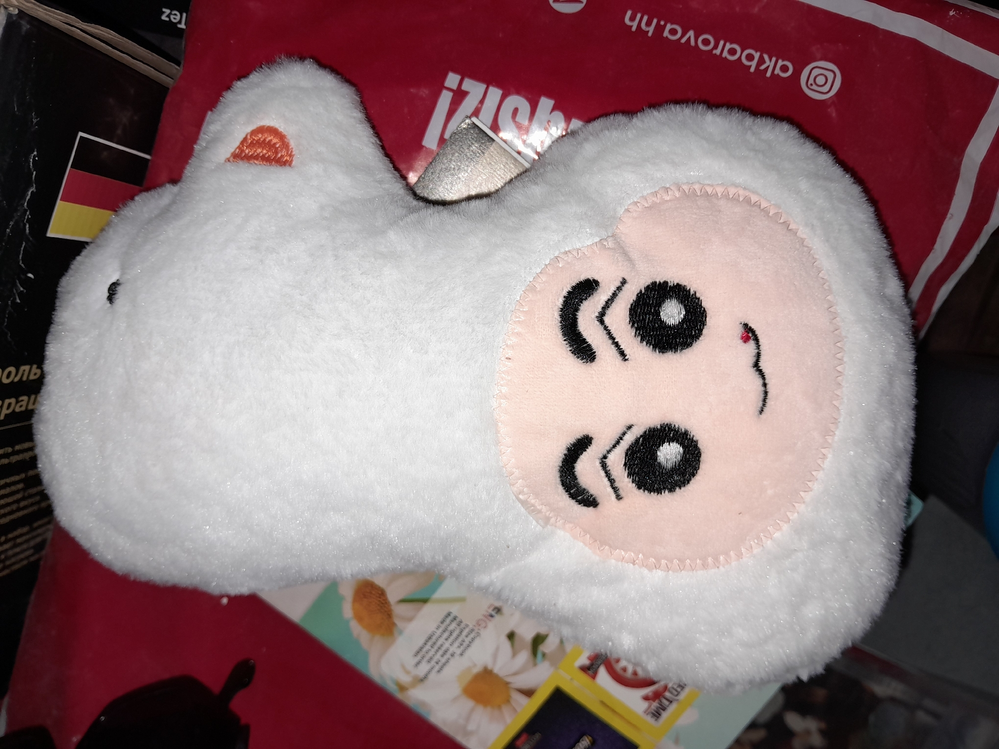 A white plush toy sheep is lying on a red surface, possibly a bag or cloth. The sheep has a pink face with embroidered black eyes, eyebrows, and a small red mouth. Its ears are orange. In the background, there are various objects including what appears to be a stack of books or boxes with text and images, and a part of a German flag. The overall setting appears to be indoors. There are no people visible in the image. The location in the city of Uchquduq, Uzbekistan, is not discernable from the visual information provided in the image.