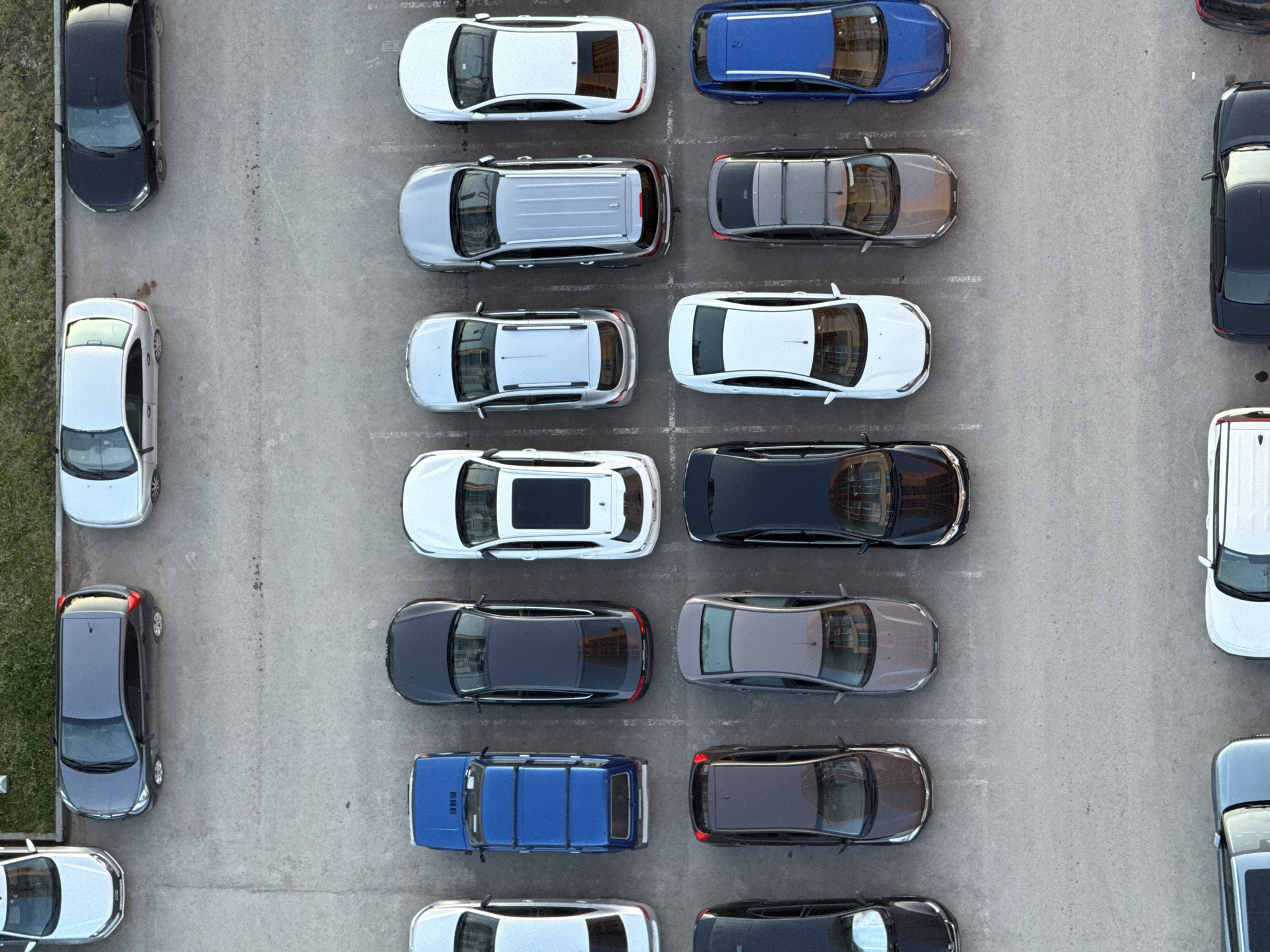 This aerial, top-down view captures a busy outdoor parking lot in Dikaya Gar', Russia. The scene is dominated by numerous parked automobiles, arranged in rows across a paved surface, likely asphalt or concrete.
**Subjects:** The primary subjects are approximately 25 to 30 cars of various makes, models, and colors. These include sedans, SUVs, and potentially hatchbacks. A wide range of colors is visible, with many cars being white, silver, light grey, dark grey, black, or dark blue. On the far left side of the image, a narrow strip of green grass and some bare earth is present, bordering the parking area. No people are visible in the frame.
**Activity or event taking place:** The image depicts a static scene of vehicles parked in a lot. There is no discernible activity or event taking place; all cars are stationary.
**Notable details:** The photograph was taken from an elevated perspective, offering a clear overhead view of the parking arrangement. The lighting indicates it is daytime, with relatively soft and even illumination, suggesting either an overcast sky or a high sun position. The weather appears clear and dry. The ground surface shows some minor imperfections and faint markings that might be remnants of parking space delineations. No specific location cues like buildings or prominent landmarks are visible beyond the cars and the ground. There is no visible text on any of the vehicles or the paved surface. The overall impression is that of a functional, well-utilized parking facility.
