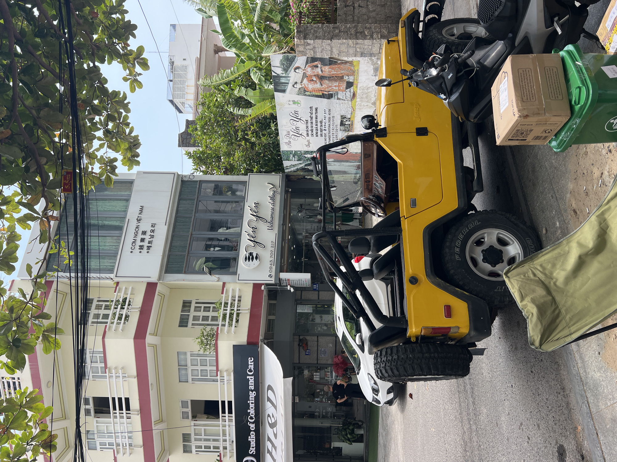 The image captures a vibrant street scene in Bắc Nha Trang, Vietnam, under a bright, clear sky, indicative of daytime. A prominent yellow open-top vehicle, resembling a classic Jeep and marked with "Jeep 1979" on its dashboard and "Jeep" on its side, is parked curbside on the right. A scooter is partially visible behind it, next to a green trash can topped with a cardboard box labeled "TOP GIA KHĂN GIẤY CAO CẤP". An olive green camping chair is collapsed beside the yellow vehicle. The tire of the yellow vehicle displays "FEDERAL".
Across the street, several multi-story buildings house various businesses. One sign prominently reads "CƠM NGON VIỆT NAM" (Vietnamese cuisine), accompanied by Korean text "베트남요리". Below it, "5.5m" is visible. Further down, "Studio of Coloring and Care H&D" operates under a white awning, next to a street number "13". Another significant storefront is "Yến Yến Vietnamese clothing", advertising "Ao Dài Yến Yến". A large poster affixed to a wall behind the yellow vehicle elaborates on "BÁN VÀ CHO THUÊ CÁC MẪU ÁO DÀI THIẾT KẾ CƯỚI, BƯNG QUẢ, BÀ SUI NHA TRANG, DÂY, VEST DẠ HỘI" (selling and renting wedding Ao Dai designs, bridal trays, bridesmaid dresses in Nha Trang, belts, evening dresses), along with the phone number "0943.707.217" and Korean text detailing traditional Vietnamese dresses.
Lush green trees with visible power lines frame the upper left of the image, and banana plants add to the tropical foliage near the buildings. A white car is parked in front of the yellow vehicle, with a person standing nearby. The overall scene conveys the bustling atmosphere of a commercial street.