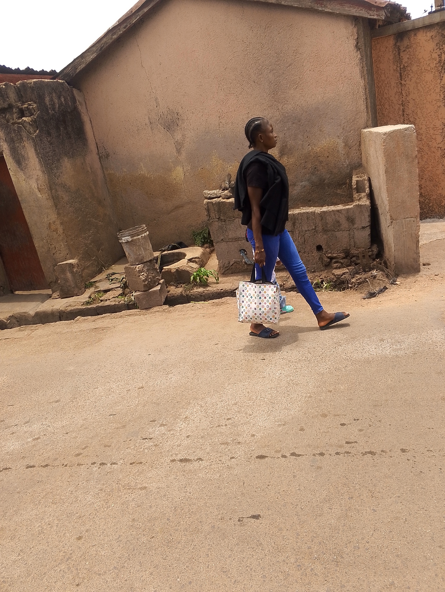 The image depicts an outdoor street scene in Jos, Nigeria, during the daytime. The primary subject is a woman, viewed in profile, walking from right to left across the frame. She has braided hair styled in cornrows, and is wearing a black top or shawl, blue jeans, and dark blue slip-on sandals. She carries a light-colored handbag adorned with a star or flower pattern in her right hand.
Behind her, to the left, a portion of another person wearing light blue sandals is visible, suggesting they might be walking together or closely behind. The road in the foreground and midground is a light brown, coarse-textured surface, possibly asphalt or compacted earth, with some darker stains or marks.
The background features weathered buildings with textured, earthy-toned walls, indicative of local construction styles. To the left, a dark wooden door is set into a distressed wall. Along the side of the road, various objects are present, including stacked concrete blocks, a metal bucket, and general rubble, with small green plants growing amidst the debris. A prominent, light-colored concrete pillar stands upright against a textured wall on the right side of the image. The sunlight suggests a clear day. No visible text is discernable on any objects or surfaces.