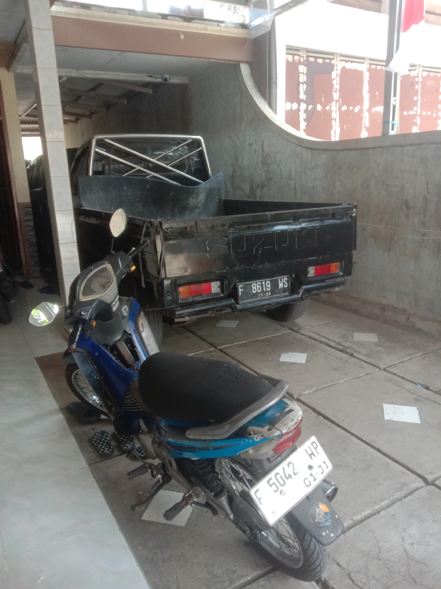 In Bojong, Indonesia, a black Suzuki pickup truck is parked under a covered area, its bed empty and a chrome roll bar visible. To the left of the pickup, a blue and black motorcycle is parked. The motorcycle has a black seat and a white license plate that reads "F 5042 HP G1.31". Another white license plate on the pickup truck reads "F 8619 WS 11-26". The ground is paved with concrete tiles, and there are a few white pieces of paper scattered on the ground. A wall with a textured finish is visible in the background, and the area appears to be a residential or commercial garage. There are no people visible in the image.
