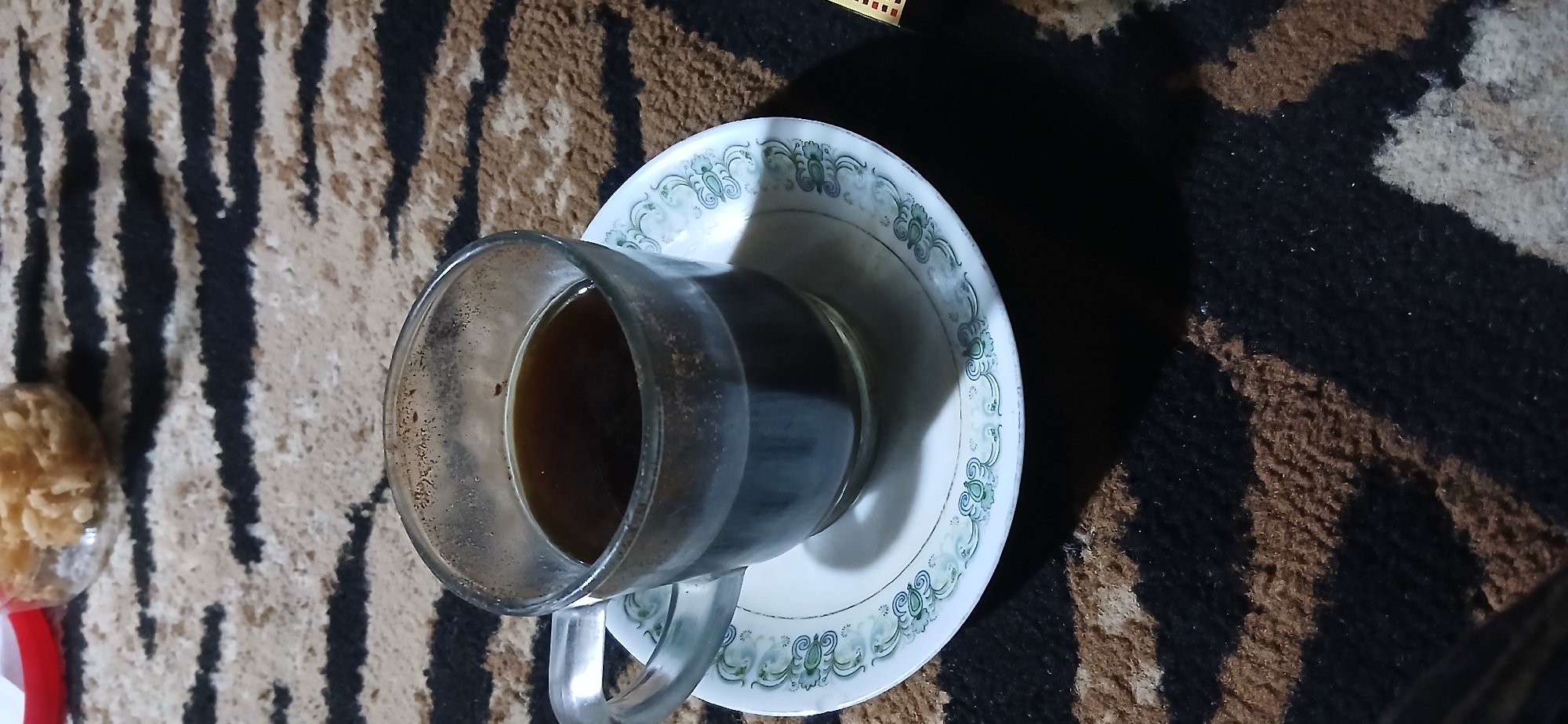 A close-up shot captures a clear glass mug filled with dark coffee, placed on a white saucer with a delicate green floral pattern. The mug and saucer rest on a carpet with a distinctive black and tan striped pattern, reminiscent of animal print. In the blurred background, a clear plastic container filled with a light-colored, crunchy snack is partially visible, suggesting a casual setting, possibly a home or small shop. The overall lighting is dim, casting a slight shadow from the mug and saucer onto the carpet, creating a cozy ambiance. The location is identified as Demangan, Indonesia.
