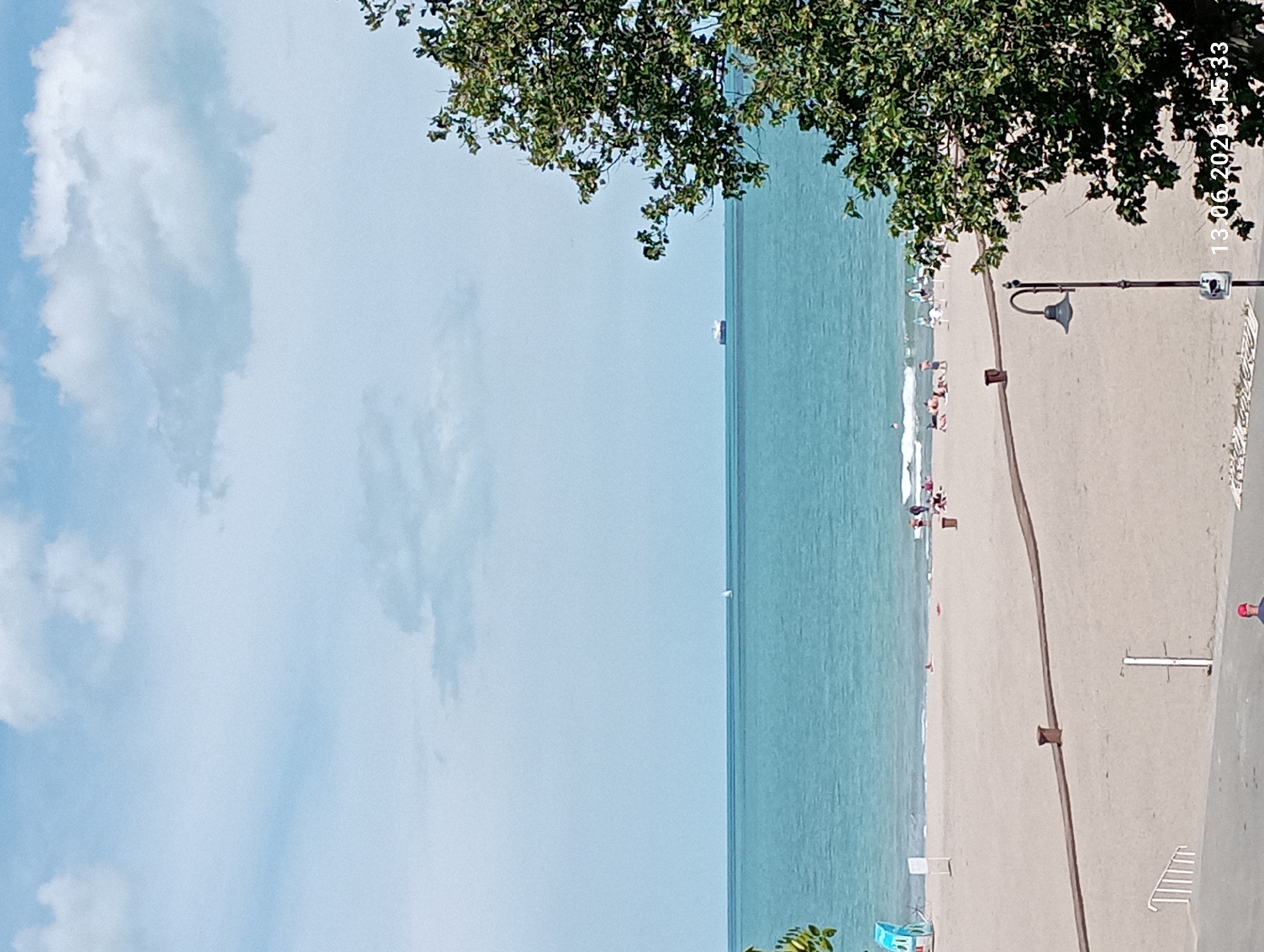 The image shows a wide view of a beach and the sea on a sunny day. In the foreground, a sandy beach stretches towards the turquoise water. Several people are scattered on the beach, some sitting or lying down, while others are closer to the water. Beach chairs and umbrellas are visible, indicating a typical beach scene.
Further out in the sea, a small sailboat can be seen, and in the distance, a large cargo ship is sailing. The sky is blue with scattered white clouds. On the right side of the image, a large, leafy green tree is prominently featured, obscuring a portion of the view. The bottom right corner of the image shows a timestamp: "13.06.2026 15:33".
This scene is consistent with the beaches found along the coast of Burgas, Bulgaria, specifically the Central Beach or the North Beach, which are popular recreational areas. The presence of the sea, sandy beach, and amenities like beach chairs are characteristic of these locations. The distant cargo ship is also a common sight in the Black Sea.