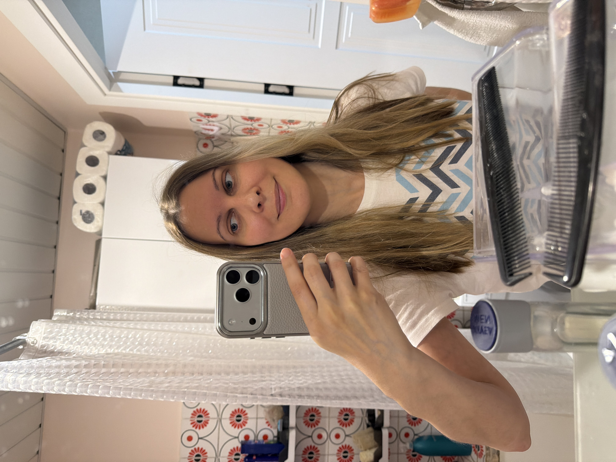 A fair-skinned woman with blonde hair and blue eyes takes a selfie in a bathroom. The woman is wearing a white t-shirt with a blue and grey chevron pattern. She is holding a grey smartphone in her left hand. The bathroom has white walls with a red floral patterned tile border. There is a roll of toilet paper visible on the left side of the frame. A shower curtain is draped in the foreground. A white cabinet is present. A clear plastic container holding a comb is in the foreground on the right. The location is not possible to determine based on the visual elements of the image alone.