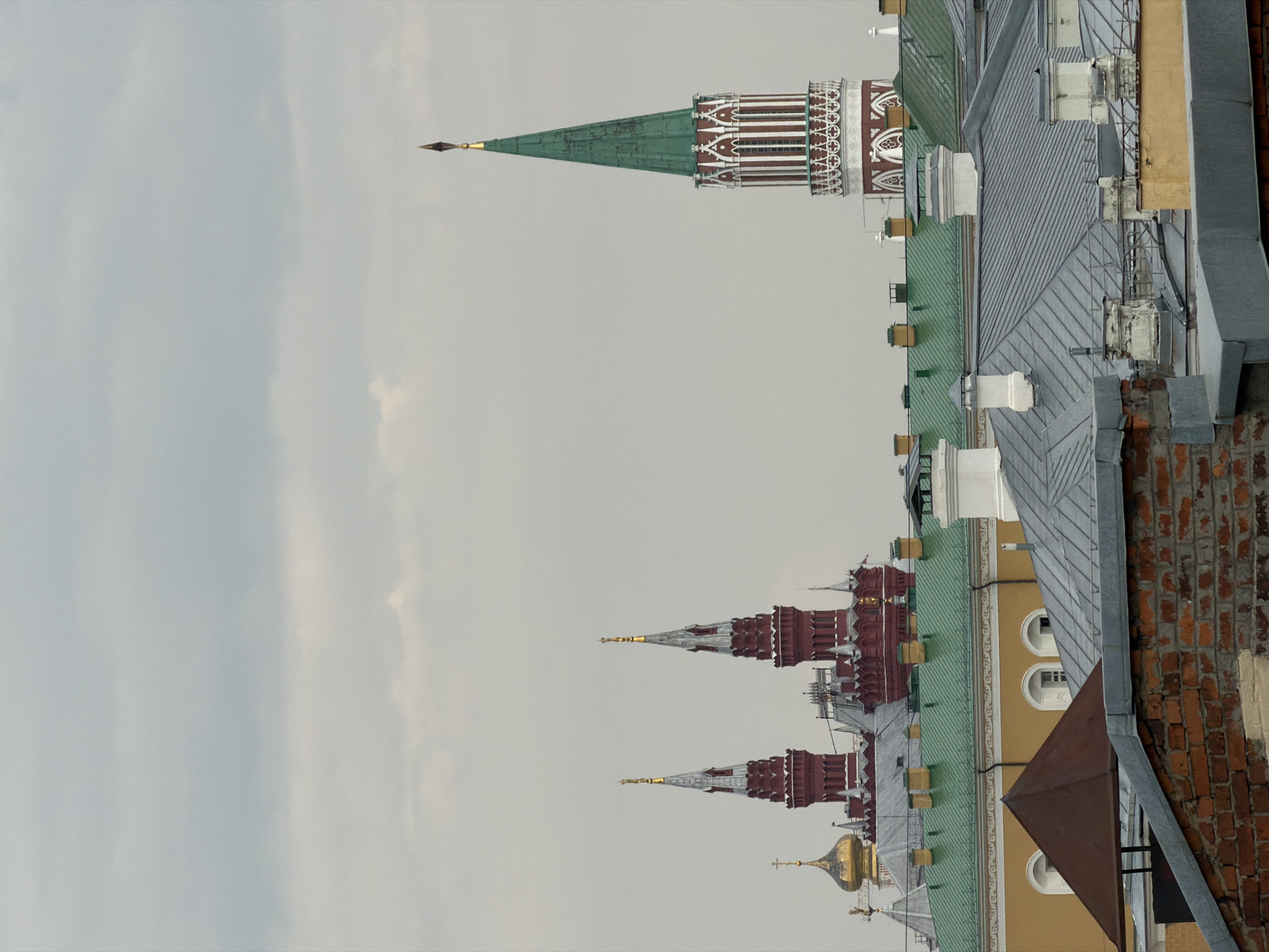 This is a vertical, medium-shot, eye-level photograph of a cityscape under a cloudy sky in Moscow, Russia. The foreground is dominated by the rooftops of various buildings, constructed with brick, grey metal, and yellow stucco. Some of these rooftops feature chimneys and skylights.
In the midground, behind the foreground rooftops, are more buildings, including a prominent yellow structure with arched windows and green-tiled roofs. Two ornate red towers with pointed spires and gold accents rise behind this building. To the right, a tall, slender tower with a green spire and white decorative elements stands out. A golden-domed building with a cross is visible to the left, behind the red towers.
The background is filled with a hazy, overcast sky. The overall impression is one of historical architecture viewed from a high vantage point. There are no people or animals visible in the frame. The image appears to be a distant view of the Moscow Kremlin and surrounding buildings.