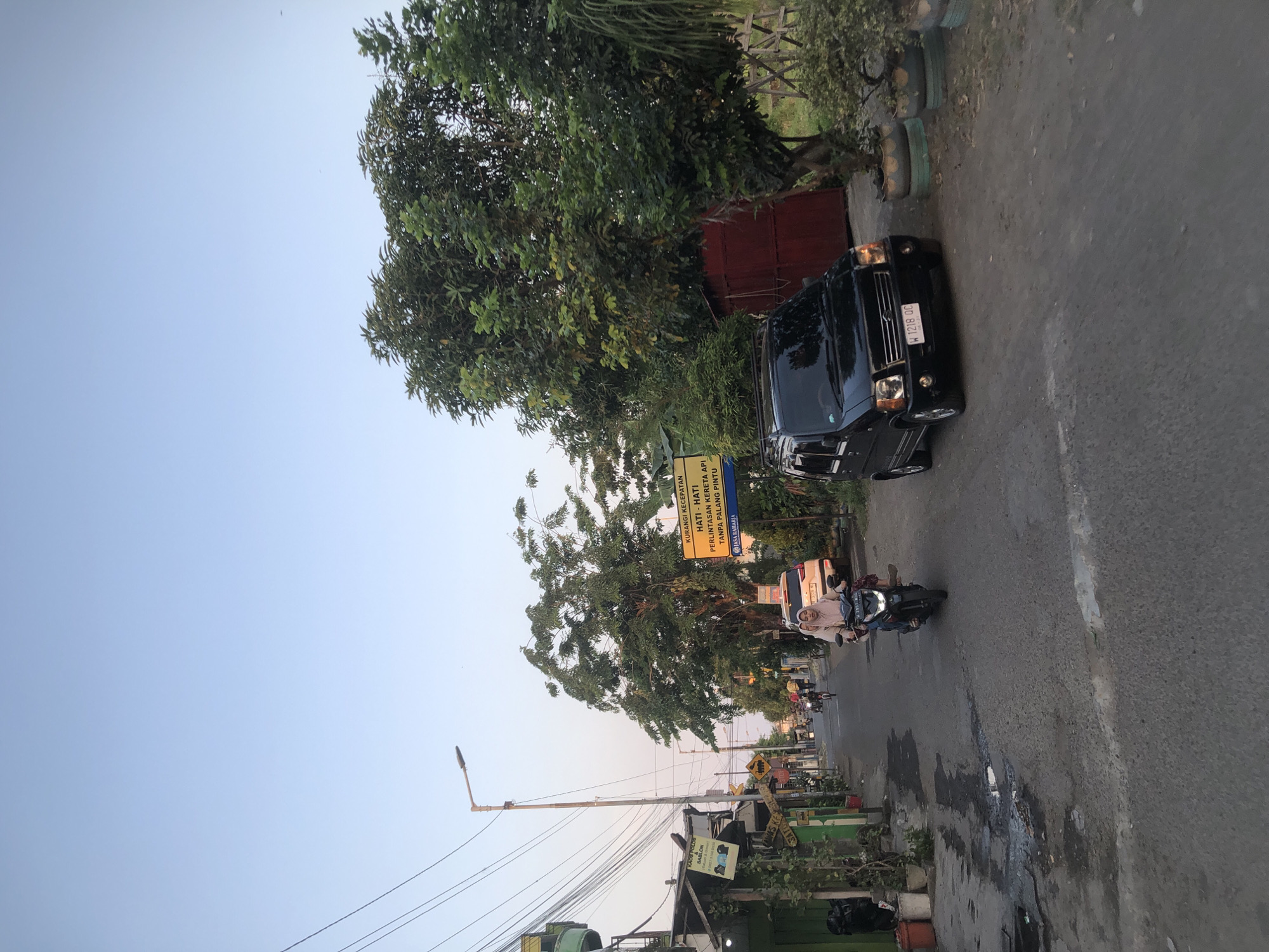 The image depicts a street scene in Tenggulunan, Indonesia, during what appears to be the late afternoon or early evening. The sky is a clear, pale blue, transitioning to a warmer hue near the horizon. Power lines are visible overhead, strung between poles.
On the right side of the street, a black Suzuki Wagon R is parked, facing forward. The car has its headlights on. Next to the car, a red structure, possibly a small shed or outbuilding, is visible, partially obscured by a large, leafy tree. Several potted plants, made from repurposed tires, are arranged around the base of the tree.
On the left side of the street, a narrow, asphalt road is lined with buildings. Several potted plants and bins are placed near the building facades. A streetlamp is positioned on a tall pole. A yellow sign with Indonesian text reading "KURANGI KECEPATAN - HATI - HATI PERLINTASAN KERETA API TANPA PALANG PINTU" (Reduce Speed - Be Careful of Unattended Train Crossings) is prominently displayed on the right side of the road. Another smaller, rectangular sign is attached to a pole on the left side of the road, also with Indonesian text.
A motorcycle is in motion on the road, carrying two individuals wearing light-colored headscarves. The driver is operating the motorcycle, and a passenger is seated behind them. Further down the road, other vehicles, including a white SUV and several motorcycles, are visible, suggesting moderate traffic. The road surface shows signs of wear and some puddles. The overall impression is of a typical residential street in a town or city in Indonesia.