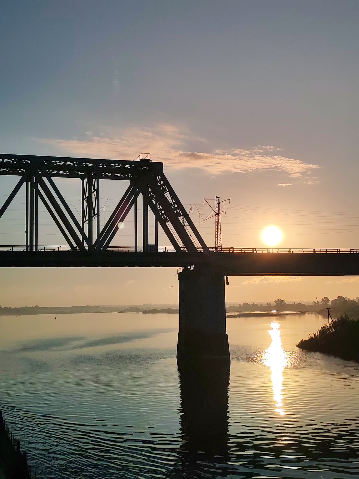 The image depicts a bridge over a body of water at sunset. The bridge is a dark silhouette against the bright sun. The sun's reflection is visible in the water. The location is Нифантово, Russia. A power line structure is also visible on the bridge.