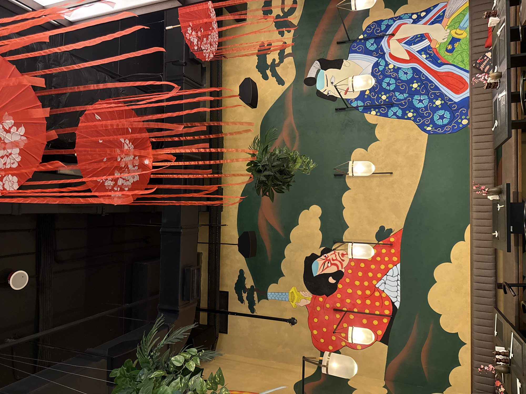 The image captures a vibrant, vertically oriented interior scene, likely within a Japanese-themed restaurant or commercial space. The central focus is a large, colorful mural painted on a high wall, depicting two stylized figures.
The upper figure in the mural is a woman wearing a blue patterned kimono, holding a fan, with a delicate, painted facial expression. Below her, another figure, a man, is portrayed in a red garment adorned with yellow dots, featuring a dramatic, mask-like face with red and white markings and a dark beard, holding a gold-hilted sword. The mural's background is characterized by flowing shapes in gold and dark green.
Above and to the left of the mural, numerous red paper umbrellas (parasols) with white cherry blossom patterns are suspended from the ceiling. They are interconnected by long, flowing red ribbons, creating a festive, layered canopy.
Throughout the decorated wall, several black metal sconces with frosted, warm-toned bulbs illuminate the mural. On the far left, dark, industrial-style architectural elements like beams and ducts are visible, along with a white circular speaker on a black surface. Lush green potted plants are placed at the bottom left and mid-left. To the far right, the edge of a dining area is visible, featuring upholstered seating and small tables adorned with delicate cherry blossom decorations in miniature vases.
The scene suggests an indoor environment, illuminated by artificial lighting. There is no specific activity or event taking place, but the decor creates a strong cultural and aesthetic atmosphere. No text is visible in the image.