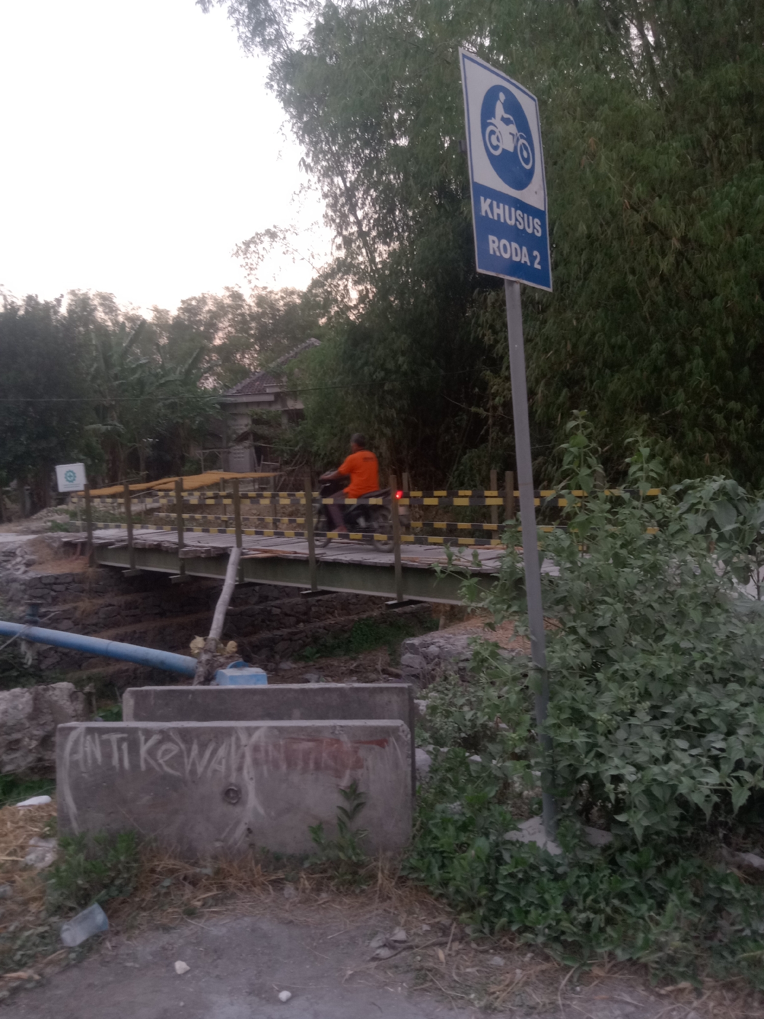 The image depicts a scene in Pacing, Indonesia, near a small bridge crossing a dry creek bed. A sign with a blue border and white background indicates "KHUSUS RODA 2", meaning "For 2 Wheels Only", with a pictogram of a motorcycle on top. The bridge itself appears to be constructed of metal beams with a wooden plank surface, bordered by yellow and black striped railings. A person on a motorcycle, wearing an orange shirt, is crossing the bridge from left to right. To the left of the bridge, a smaller white sign with a circular symbol is visible. In the foreground, concrete barriers with graffiti that reads "ANTI KEWAY" are present, along with some dry grass and a discarded plastic cup. Dense vegetation, including trees and bamboo, surrounds the area. The sky is overcast, suggesting either early morning or late afternoon.