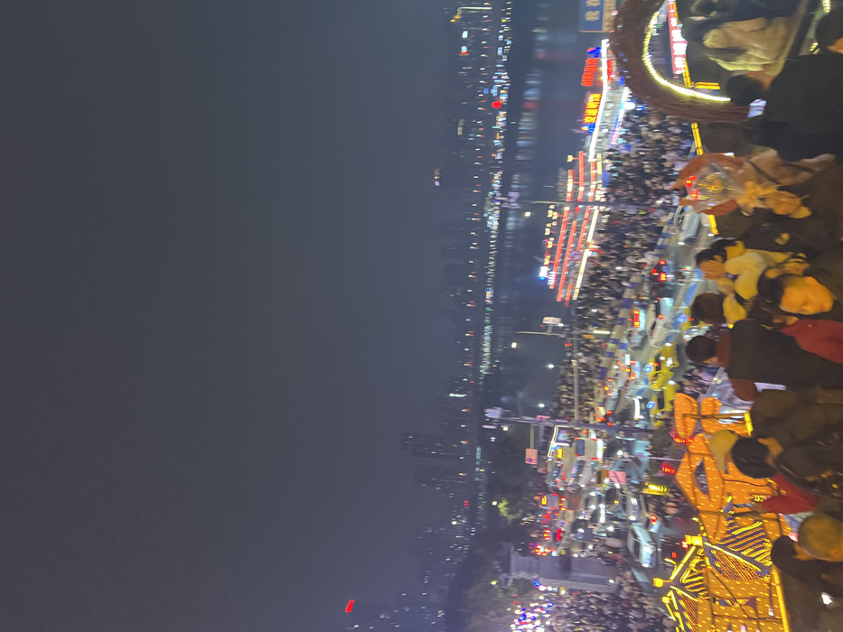 The image depicts a crowded nighttime urban landscape in Yuzhong District, China. The vantage point is elevated, looking down upon a bustling scene along a riverfront.
In the immediate foreground, a dense congregation of individuals occupies a viewing platform or walkway. The heads and upper torsos of approximately 20-30 people are visible, primarily facing away from the camera, observing the panorama below. Many are dressed in dark winter attire. Notable individuals include one person in a red jacket near the lower left, and another person holding a clear balloon adorned with ribbons, slightly right of center. Decorative structures, possibly part of an archway or railing, are visible on the right, illuminated with yellow and orange lights, featuring intricate lattice patterns.
Below this foreground crowd, a multi-lane thoroughfare is heavily congested with vehicular traffic, including numerous cars, buses, and several yellow taxis. Vehicle headlights and taillights are prominent, indicating movement or heavy idling. Adjacent to the road, another large crowd of pedestrians occupies the spaces leading down to the riverbank.
The midground features a wide river, reflecting the artificial lights of the city. A large, multi-decked passenger vessel, illuminated with prominent red and white lights, is positioned near the riverbank, possibly docked or navigating slowly. Chinese characters are visible on its side. Across the river, the background reveals a vast city skyline composed of numerous high-rise buildings, all brightly lit against the dark night sky. The city lights extend horizontally, creating reflections on the water's surface. The sky itself is dark grey, indicating complete nighttime.