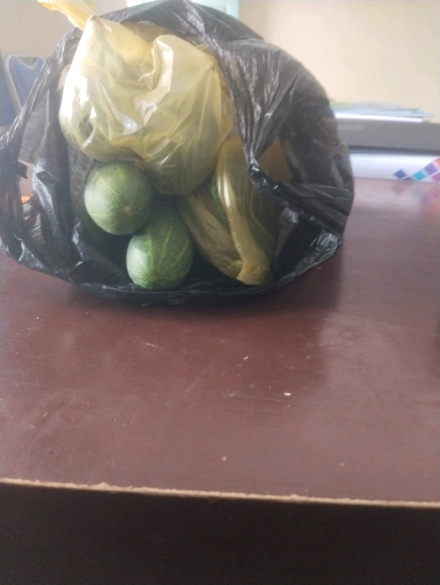A black plastic bag containing several green vegetables, possibly cucumbers or gourds, sits on a dark brown table. One of the vegetables is partially visible, with three prominent ones at the forefront of the bag. The bag is partly transparent, revealing more of the contents within. The table surface appears to be slightly textured and reflects some light. In the background, out of focus, a white object with colorful bands is visible, along with what seems to be a desk or counter. The lighting suggests an indoor environment, possibly an office or a home. No people are present in the image. The scene does not provide any specific details that can pinpoint its location to Kareng Baru, Indonesia.
