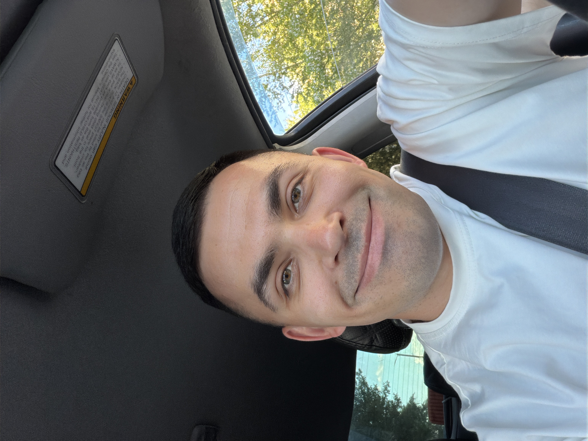 A man is depicted in a sideways, close-up selfie taken inside a car during the daytime, likely in Izhevsk, Russia. He has short, dark hair and light stubble, with brown eyes, and is wearing a white short-sleeved t-shirt. He looks directly at the camera with a slight, pleasant smile.
The car's interior is visible, including a dark dashboard and what appears to be a seatbelt across his chest. Through the car windows, lush green trees and a bright, clear sky indicate sunny weather. A prominent safety warning label is affixed to the sun visor above him.
The visible text on the warning label reads: "A WARNING DEATH OR SERIOUS INJURY can occur Children 12 and under can be killed by the air bag The BACK SEAT is the SAFEST place for children. NEVER put rear-facing child seat in the front. Sit as far back as possible from the air bag. ALWAYS Use SEAT BELTS and CHILD RESTRAINTS See the Owner's Manual for further information and explanations".