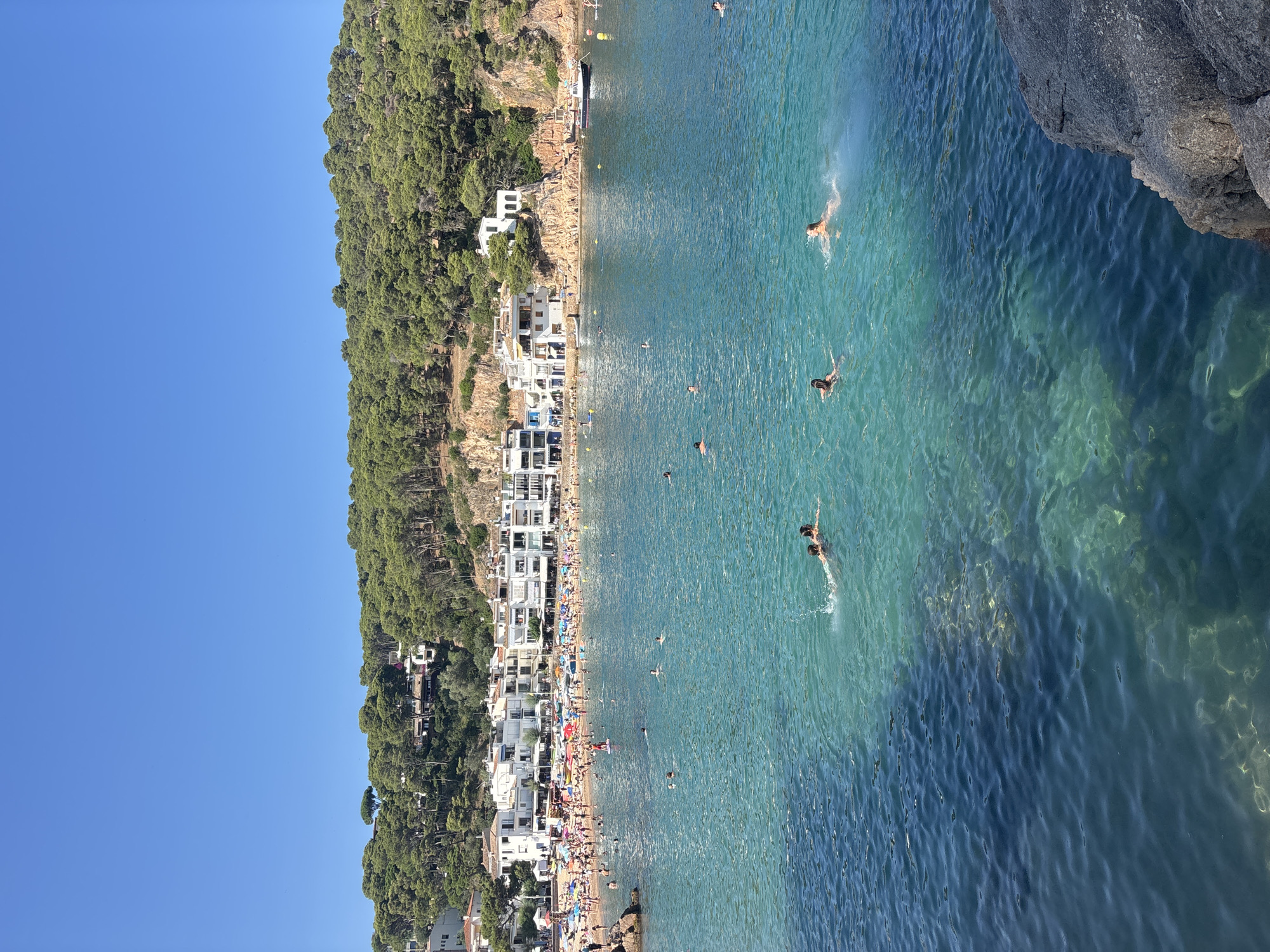 This image captures a vibrant beach scene in Calella de Palafrugell, a picturesque coastal town within the municipality of Palafrugell, Spain. The focal point is a bustling beach filled with numerous sunbathers and swimmers enjoying a clear, sunny day. The water is a striking turquoise, shallow near the shore and gradually deepening to a rich blue.
In the foreground, several individuals are swimming in the crystal-clear water. Two people are particularly visible, one with dark hair and another with lighter hair, appearing to be swimming or playing in the water. Further out, more swimmers are scattered across the sea.
The beach itself is packed with people, many lounging under colorful umbrellas. Boats are pulled up onto the sand, and a small motorboat is visible further out in the bay.
Behind the beach, a charming cluster of white-washed buildings with terracotta roofs rises up the hillside, characteristic of Mediterranean coastal architecture. These buildings are nestled amongst lush green pine trees that cover the slopes. The architecture suggests a well-established coastal village. The sky above is a brilliant, cloudless blue, indicating excellent weather conditions. The overall impression is one of a lively and idyllic summer day at a popular seaside resort.